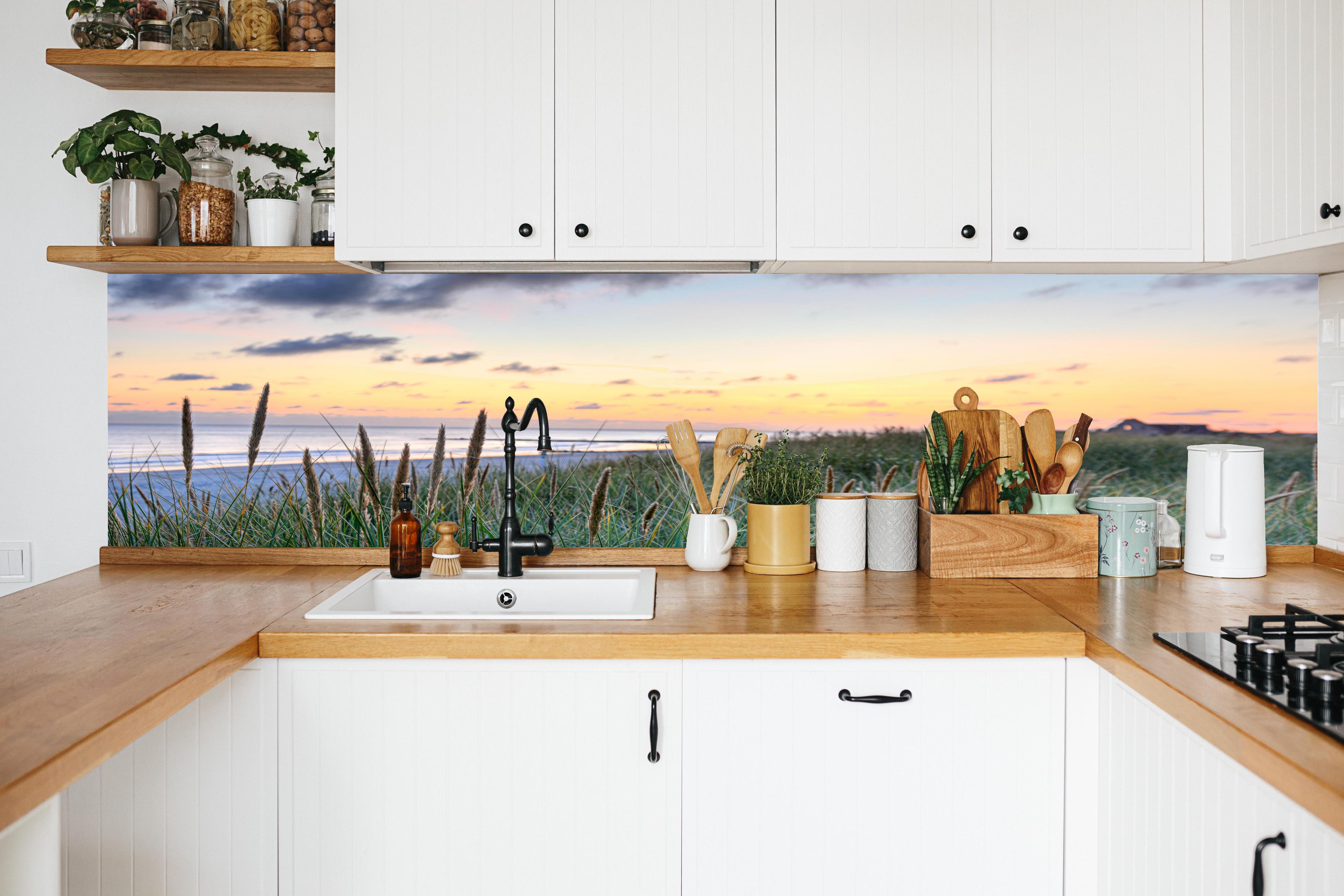 Küche - Friedliche Strandlandschaft mit grünem Gras und sanften Wellen bei Sonnenuntergang hinter weißen Hochglanz-Küchenregalen und schwarzem Wasserhahn