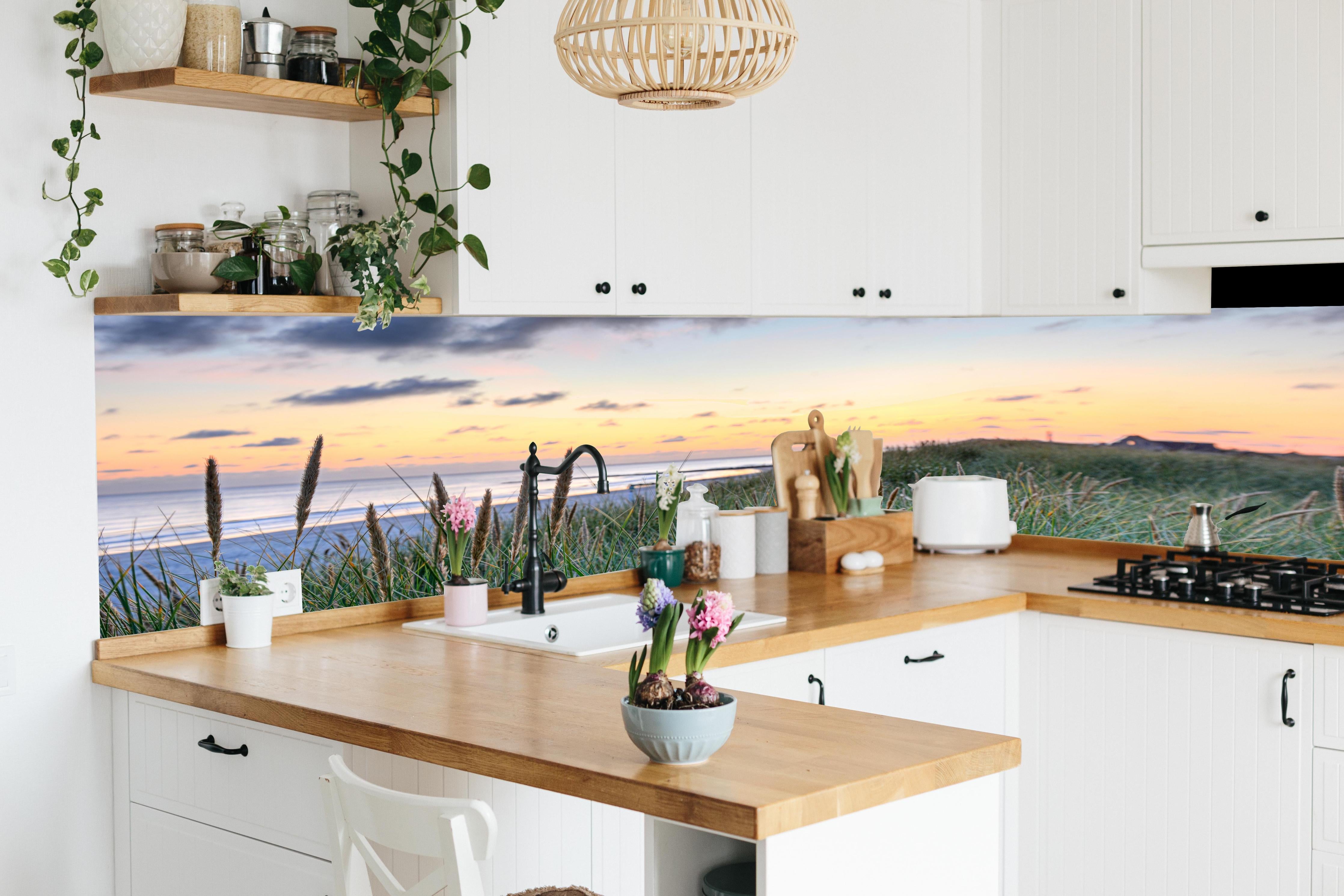 Küche - Friedliche Strandlandschaft mit grünem Gras und sanften Wellen bei Sonnenuntergang hinter weißen Hochglanz-Küchenregalen und schwarzem Wasserhahn
