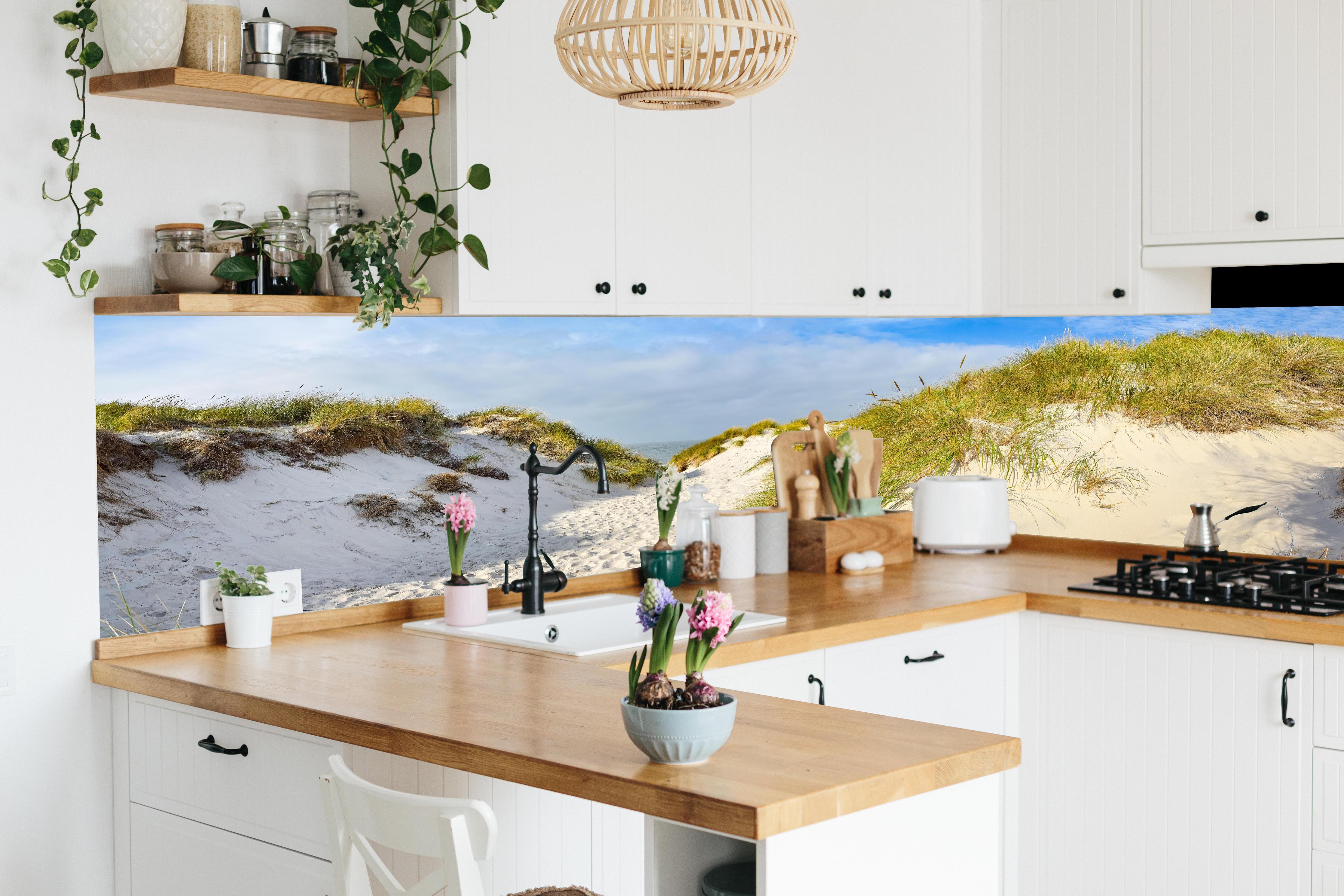 Küche - Idyllische Dünenlandschaft mit feinem Sand und grünem Gras hinter weißen Hochglanz-Küchenregalen und schwarzem Wasserhahn