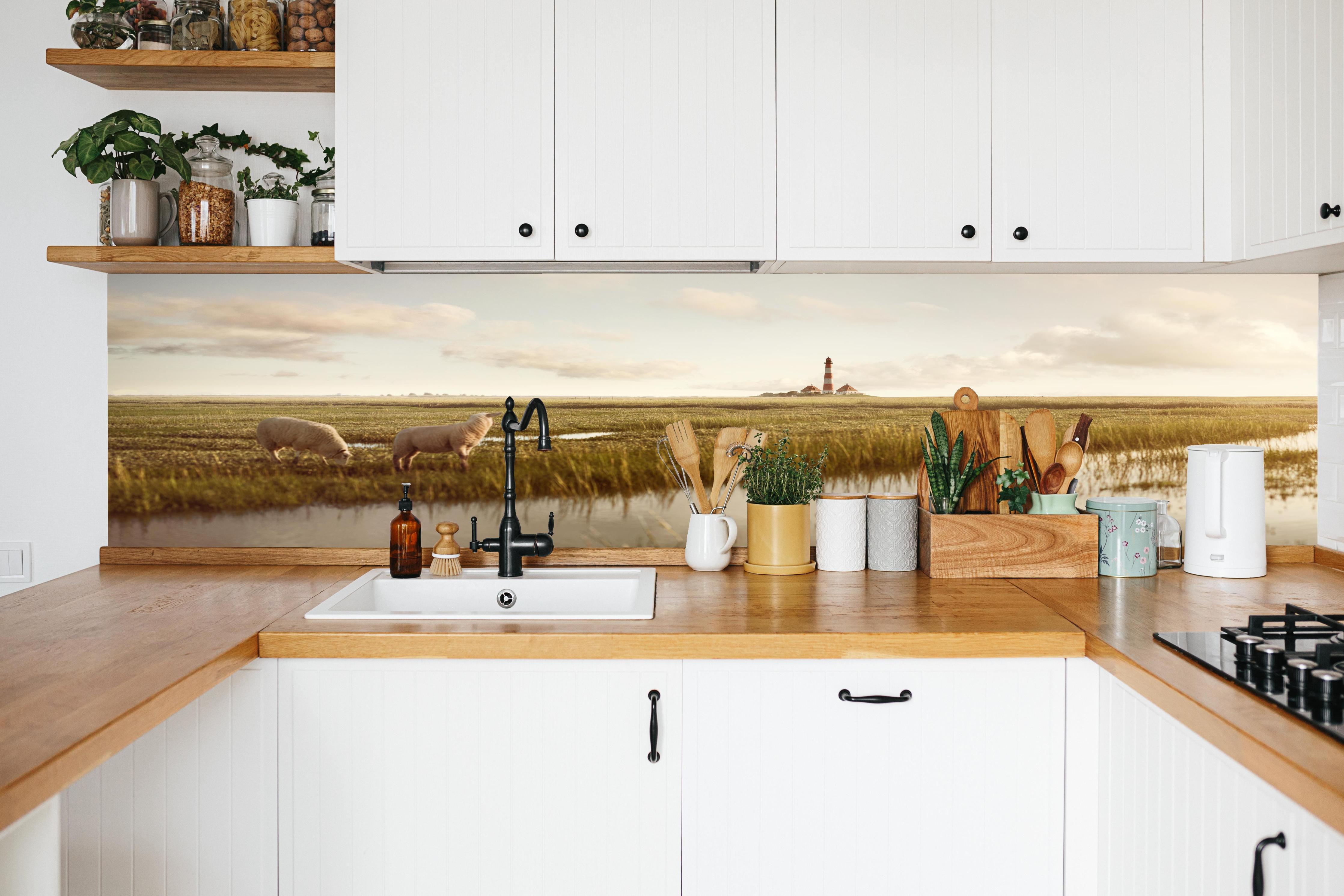 Küche - Idyllische Kulisse mit Schafen und einem Leuchtturm in ruhiger Küstenlandschaft hinter weißen Hochglanz-Küchenregalen und schwarzem Wasserhahn