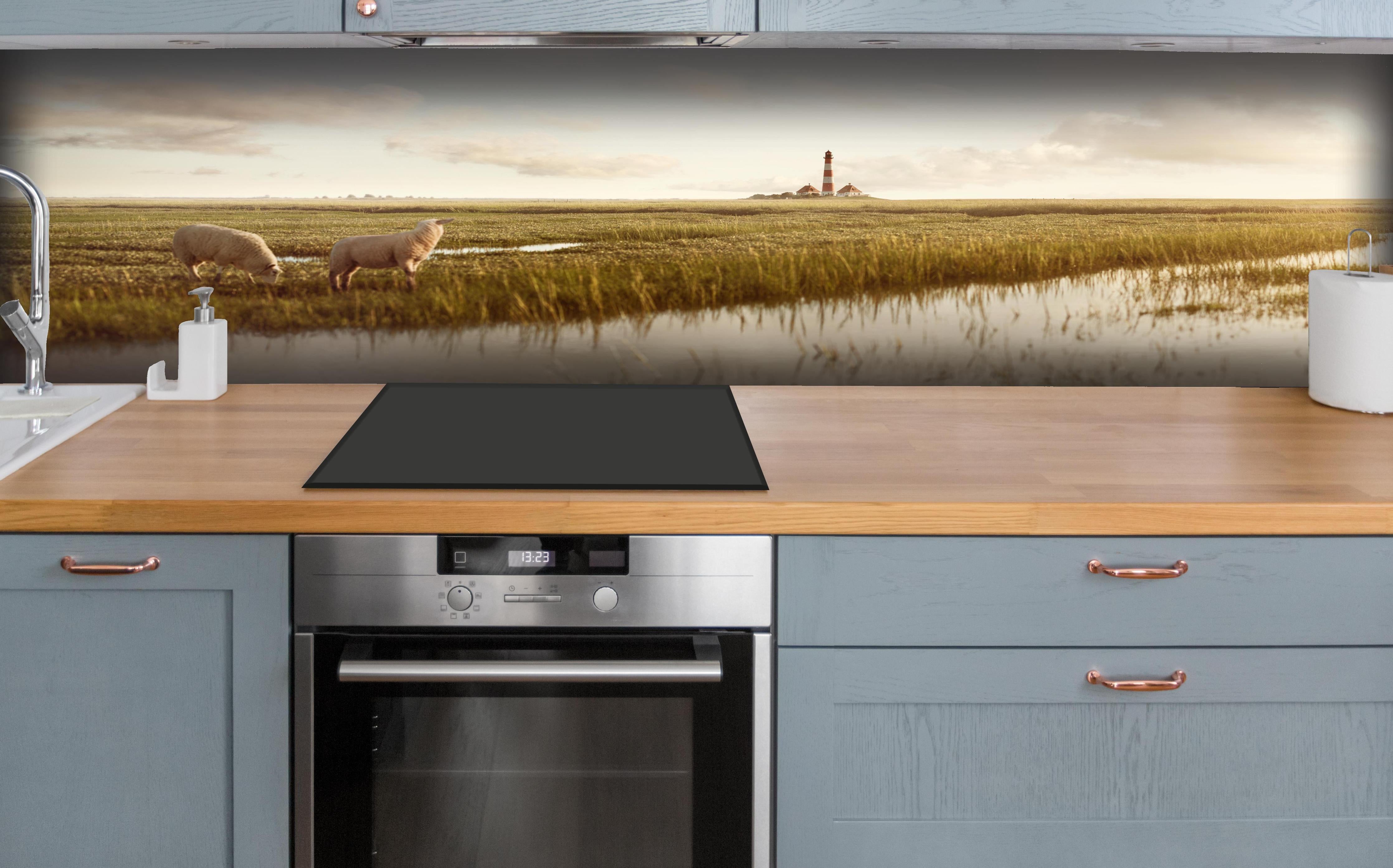 Küche - Idyllische Kulisse mit Schafen und einem Leuchtturm in ruhiger Küstenlandschaft hinter weißen Hochglanz-Küchenregalen und schwarzem Wasserhahn