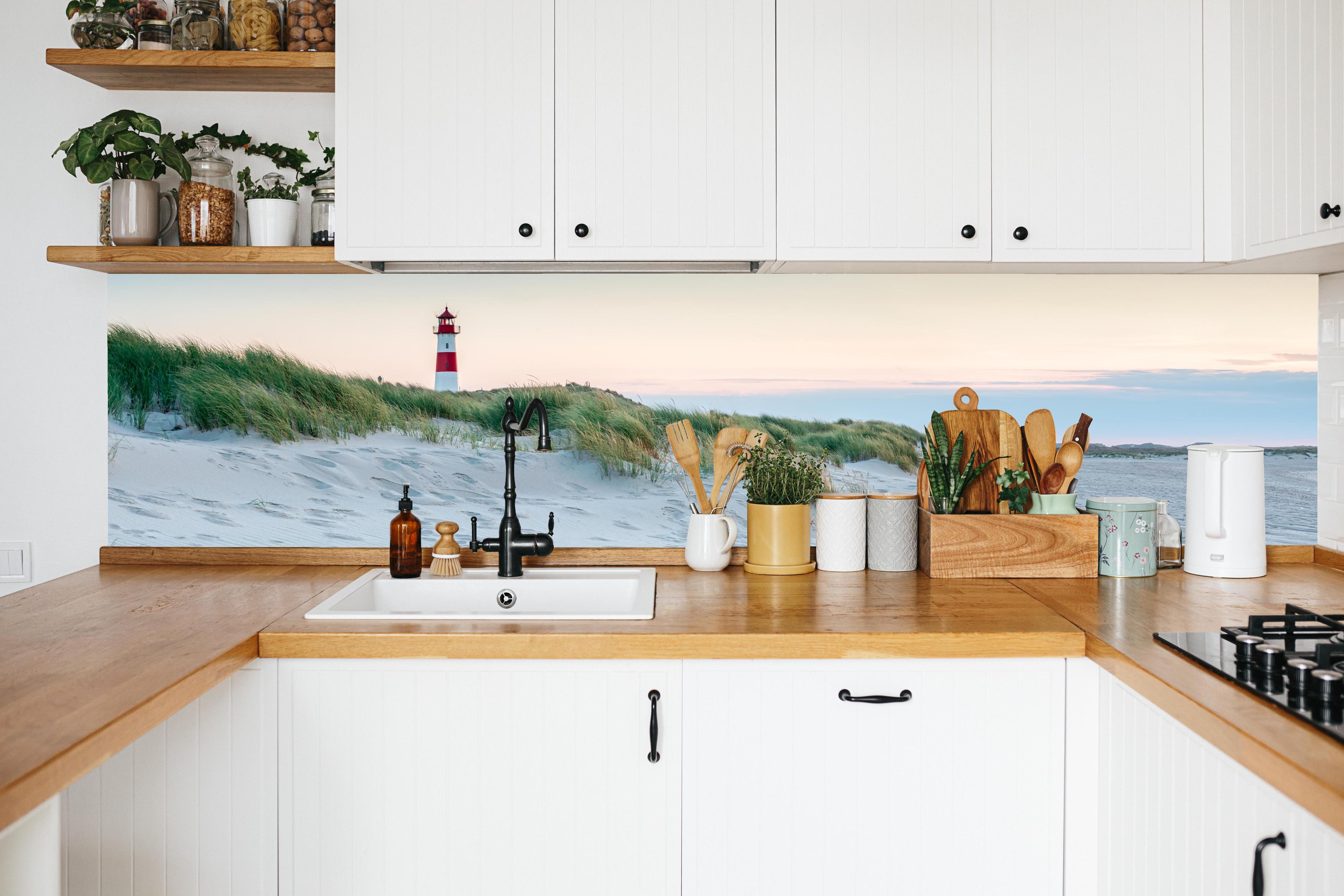 Küche - Idyllischer Leuchtturm zwischen Sanddünen und sanften Wellen hinter weißen Hochglanz-Küchenregalen und schwarzem Wasserhahn