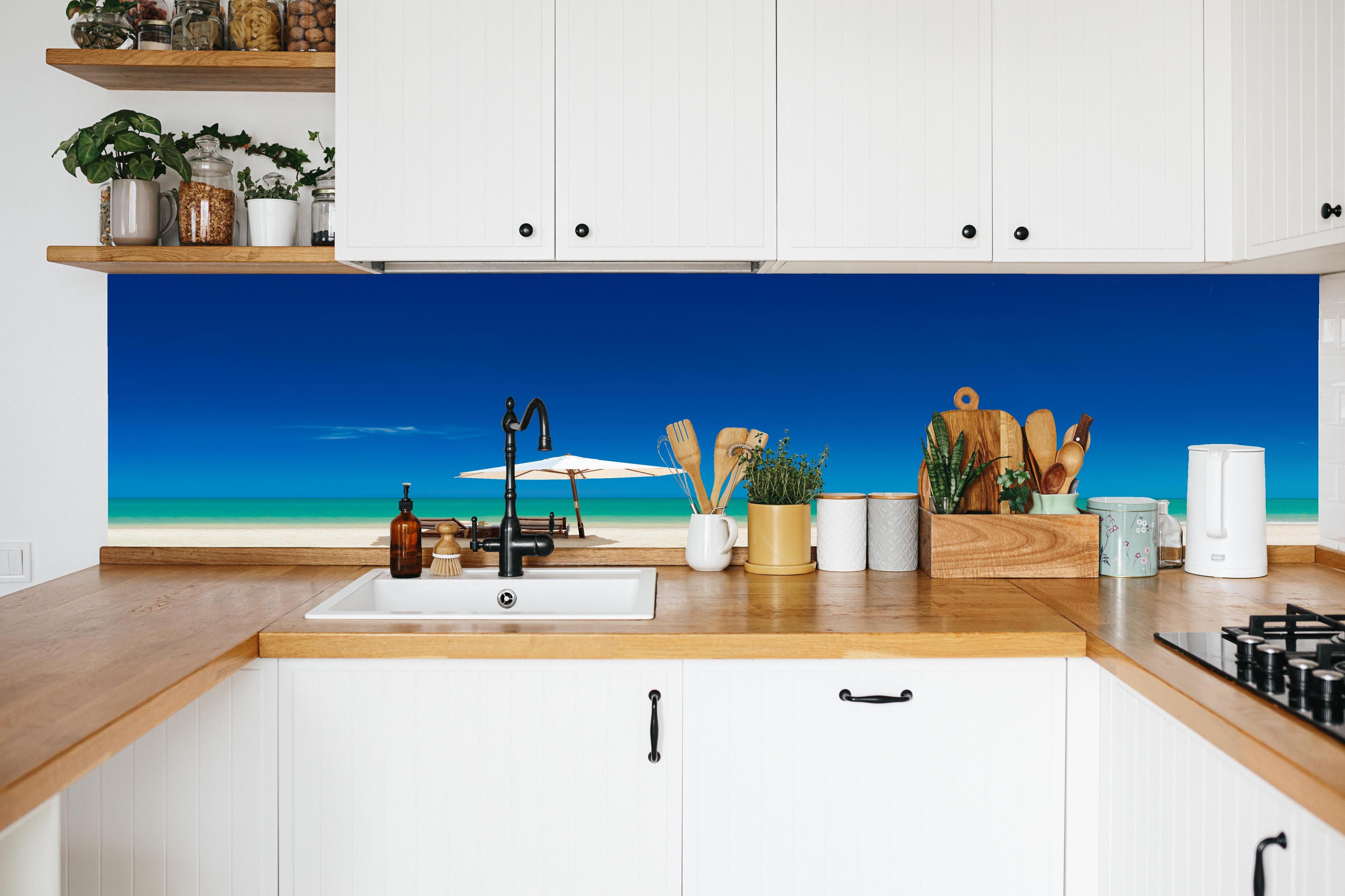 Küche - Idyllischer Strand mit Liegen und Sonnenschirm unter blauem Himmel hinter weißen Hochglanz-Küchenregalen und schwarzem Wasserhahn