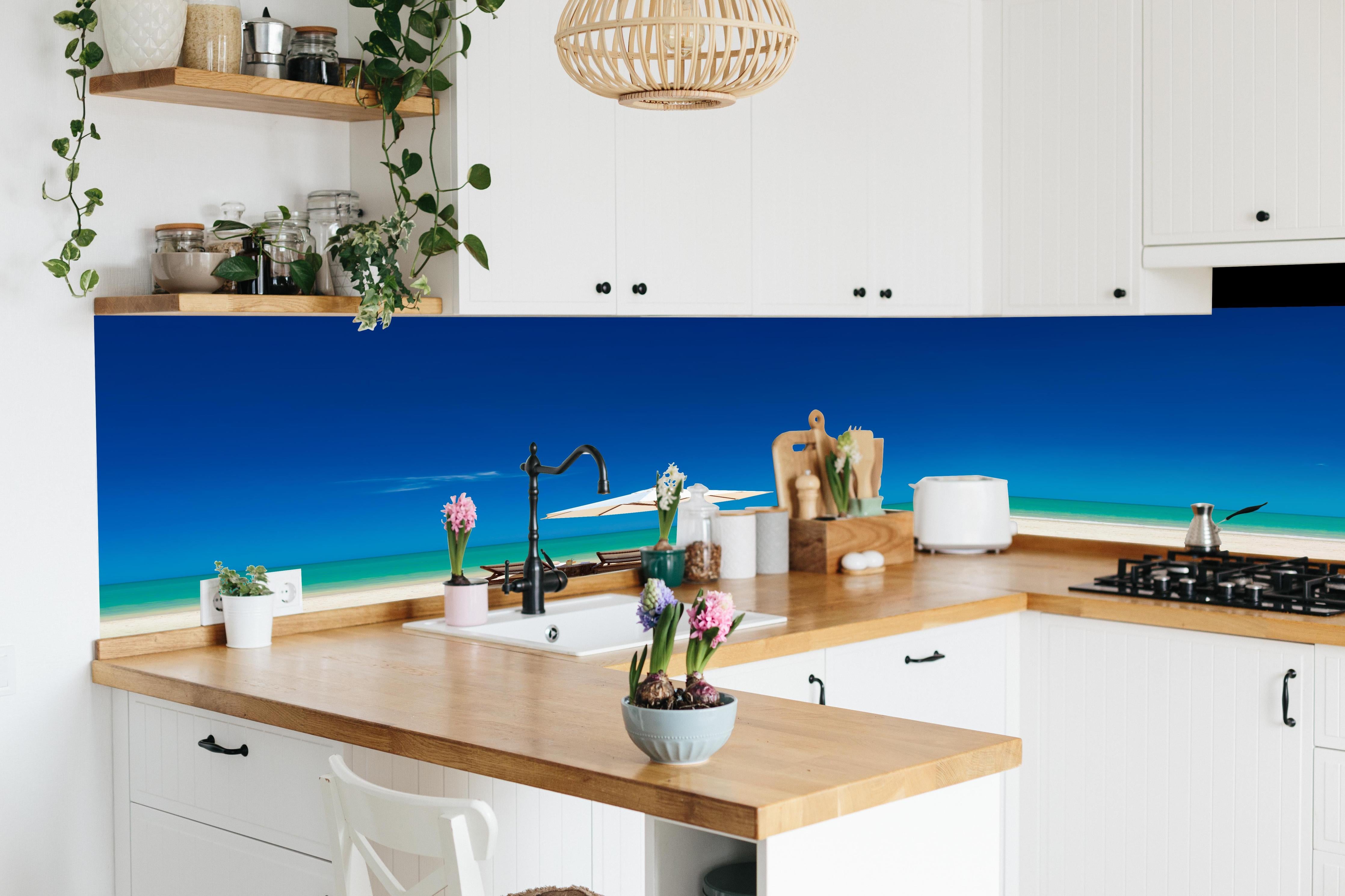 Küche - Idyllischer Strand mit Liegen und Sonnenschirm unter blauem Himmel hinter weißen Hochglanz-Küchenregalen und schwarzem Wasserhahn