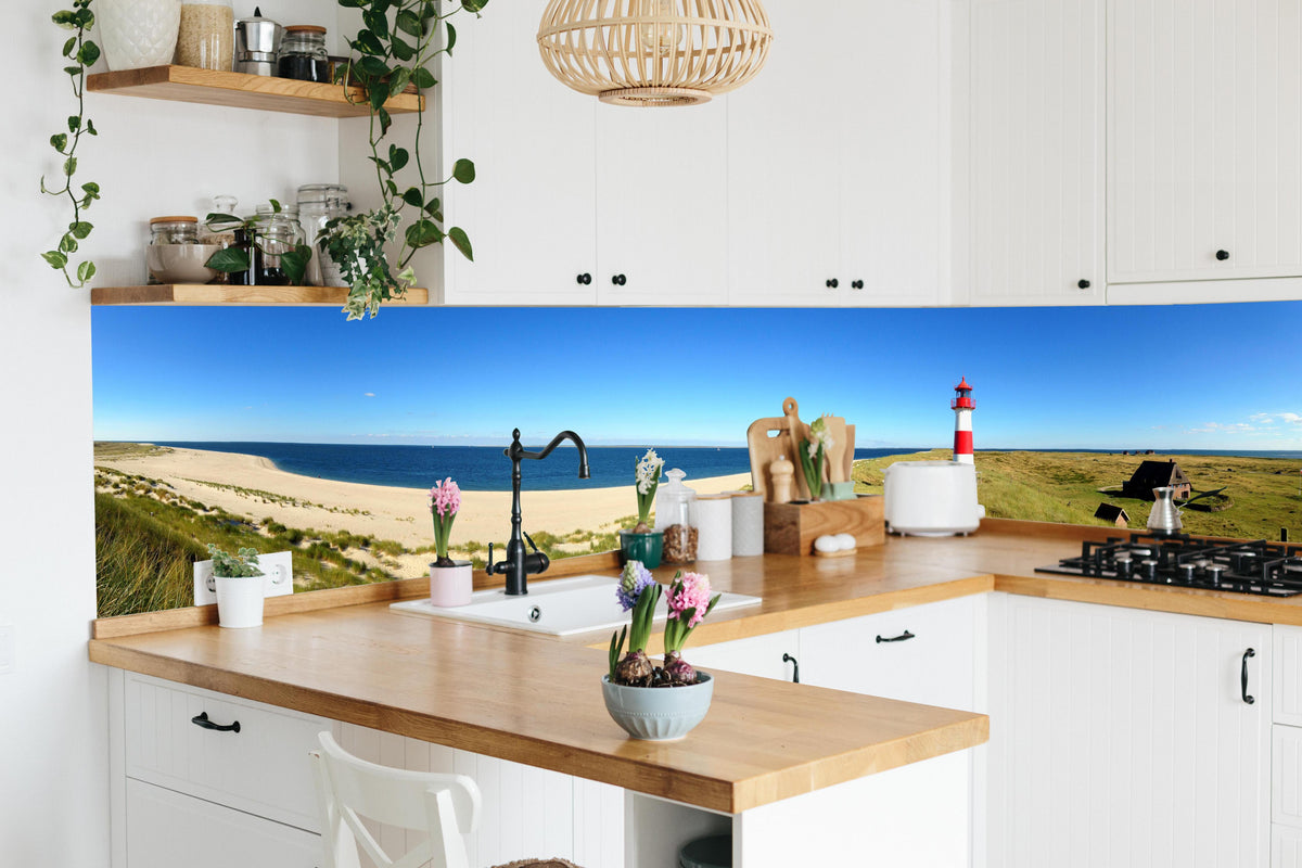 Küche - Leuchtender Leuchtturm an idyllischem Sandstrand hinter weißen Hochglanz-Küchenregalen und schwarzem Wasserhahn