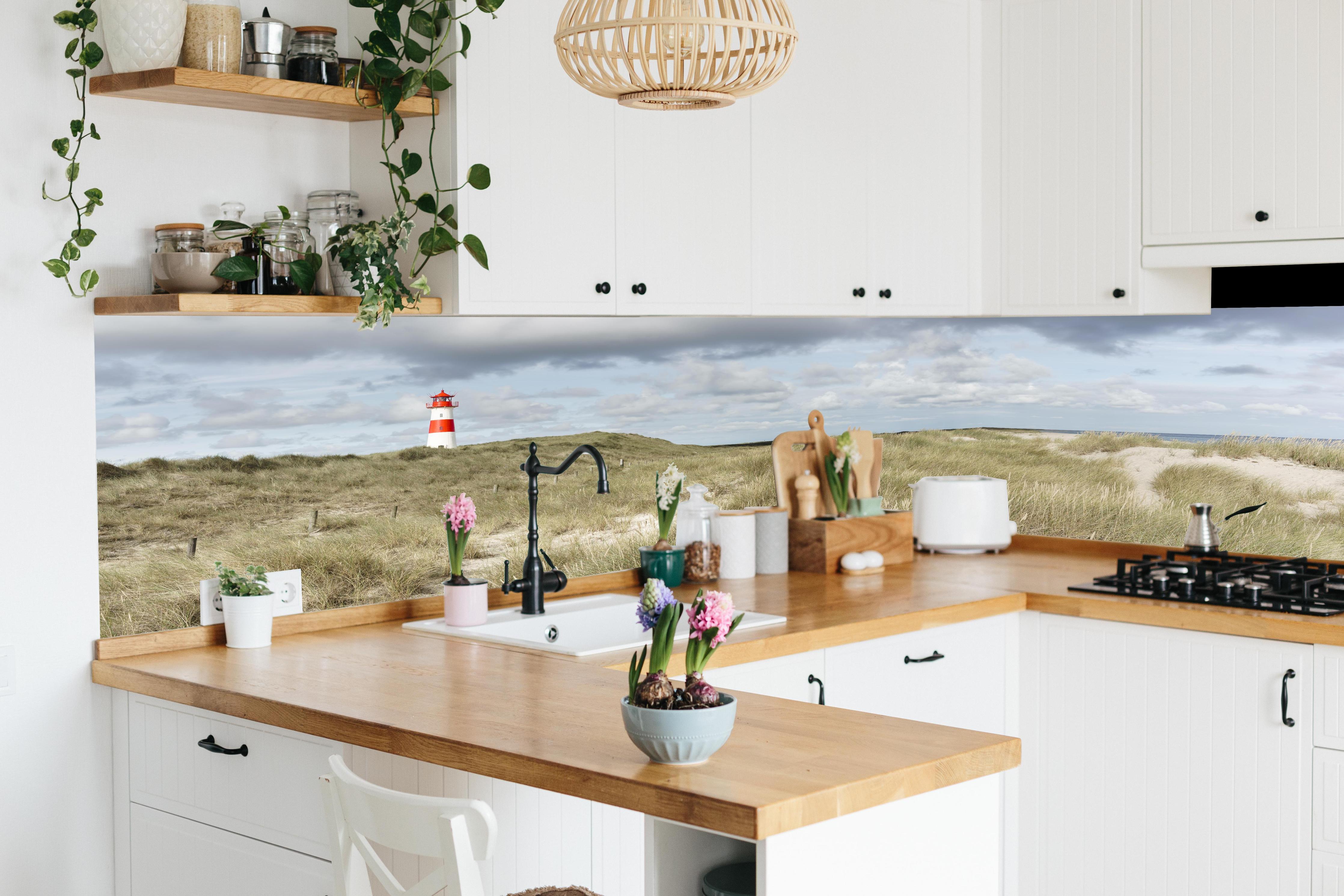 Küche - Malerischer Leuchtturm in den Dünen einer Küstenlandschaft hinter weißen Hochglanz-Küchenregalen und schwarzem Wasserhahn
