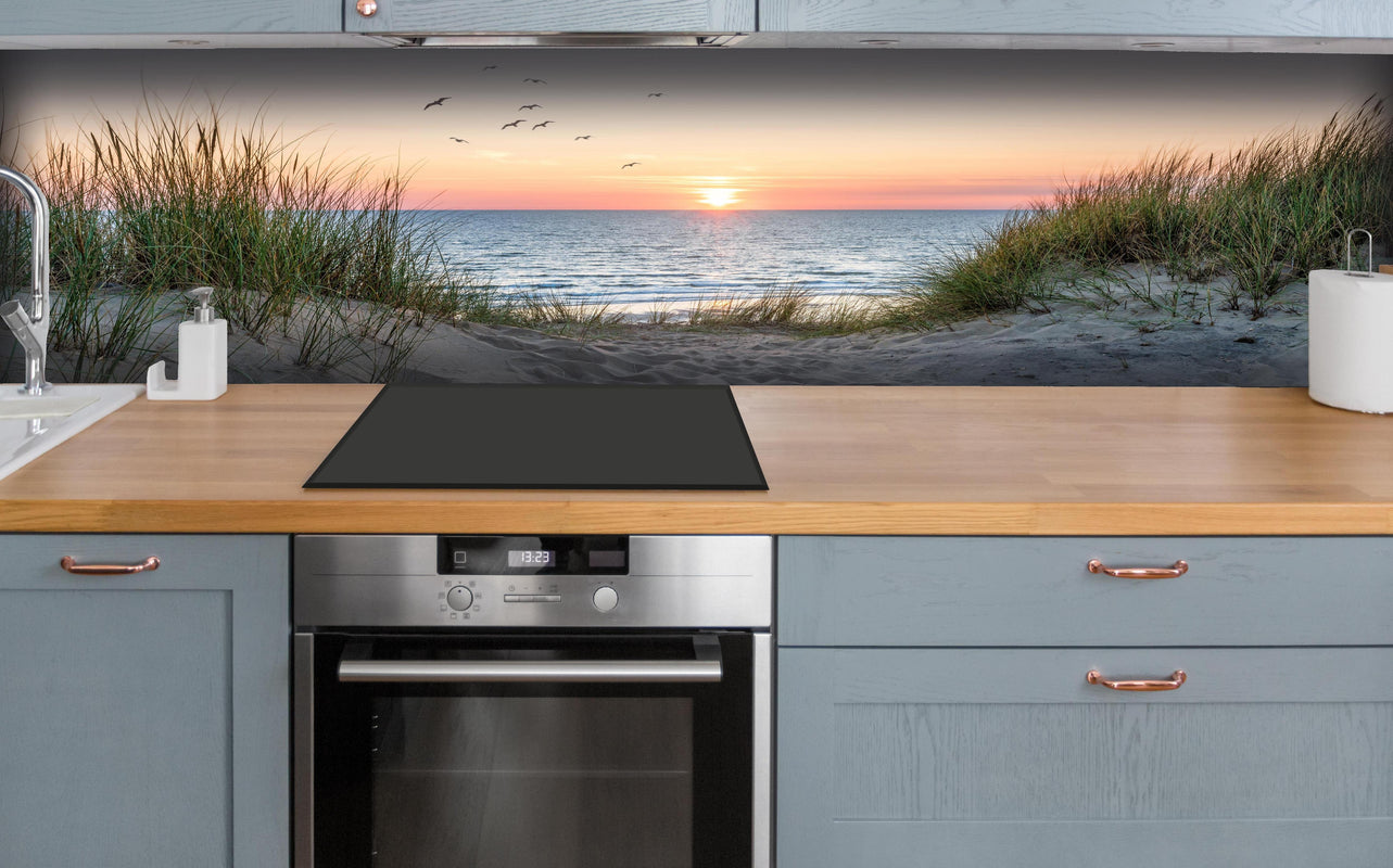 Küche - Malerischer Sonnenuntergang am Dünenstrand hinter weißen Hochglanz-Küchenregalen und schwarzem Wasserhahn