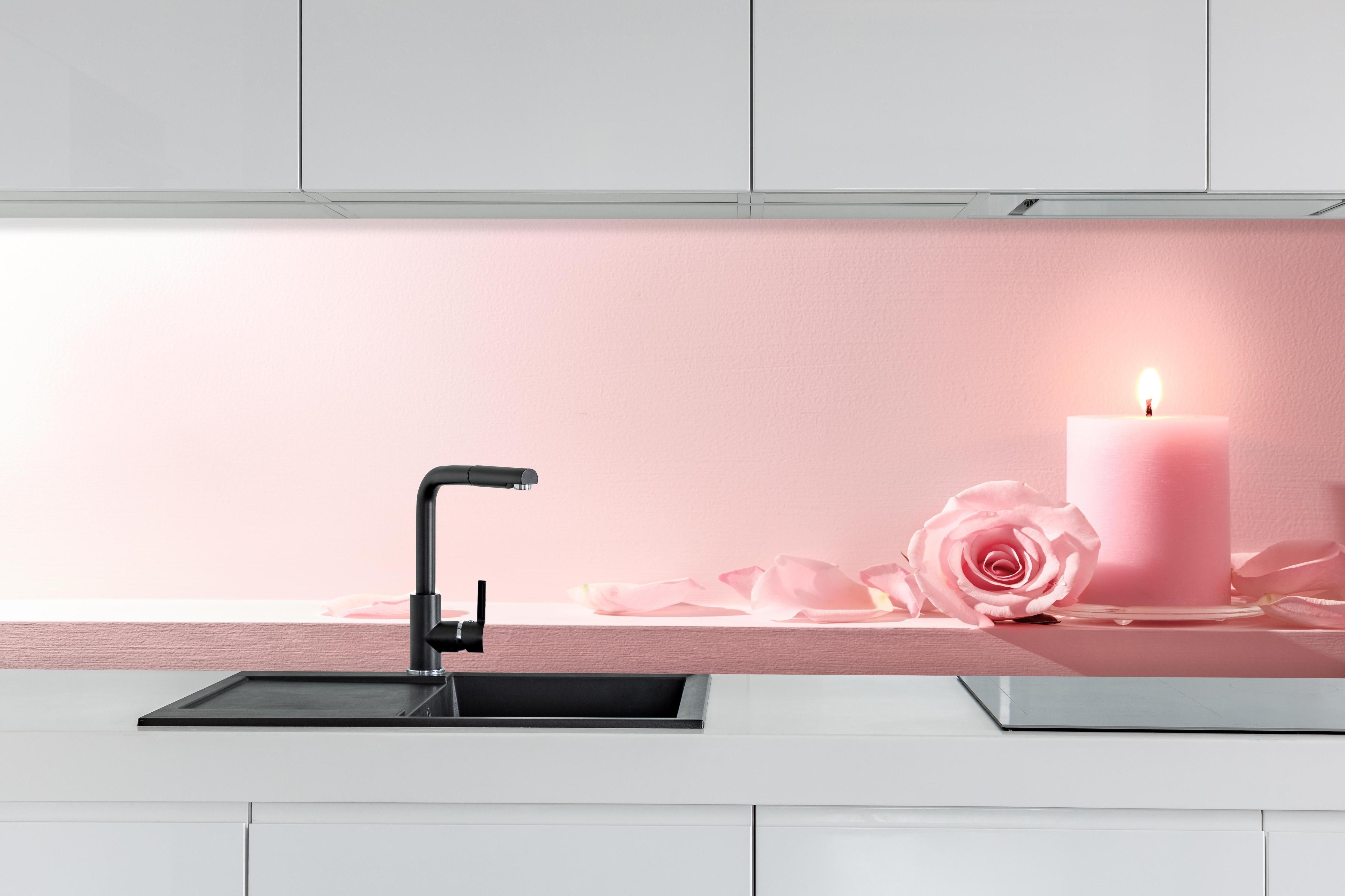 Küche - Rosa Rosen und Kerze auf einem rosa Hintergrund hinter weißen Hochglanz-Küchenregalen und schwarzem Wasserhahn