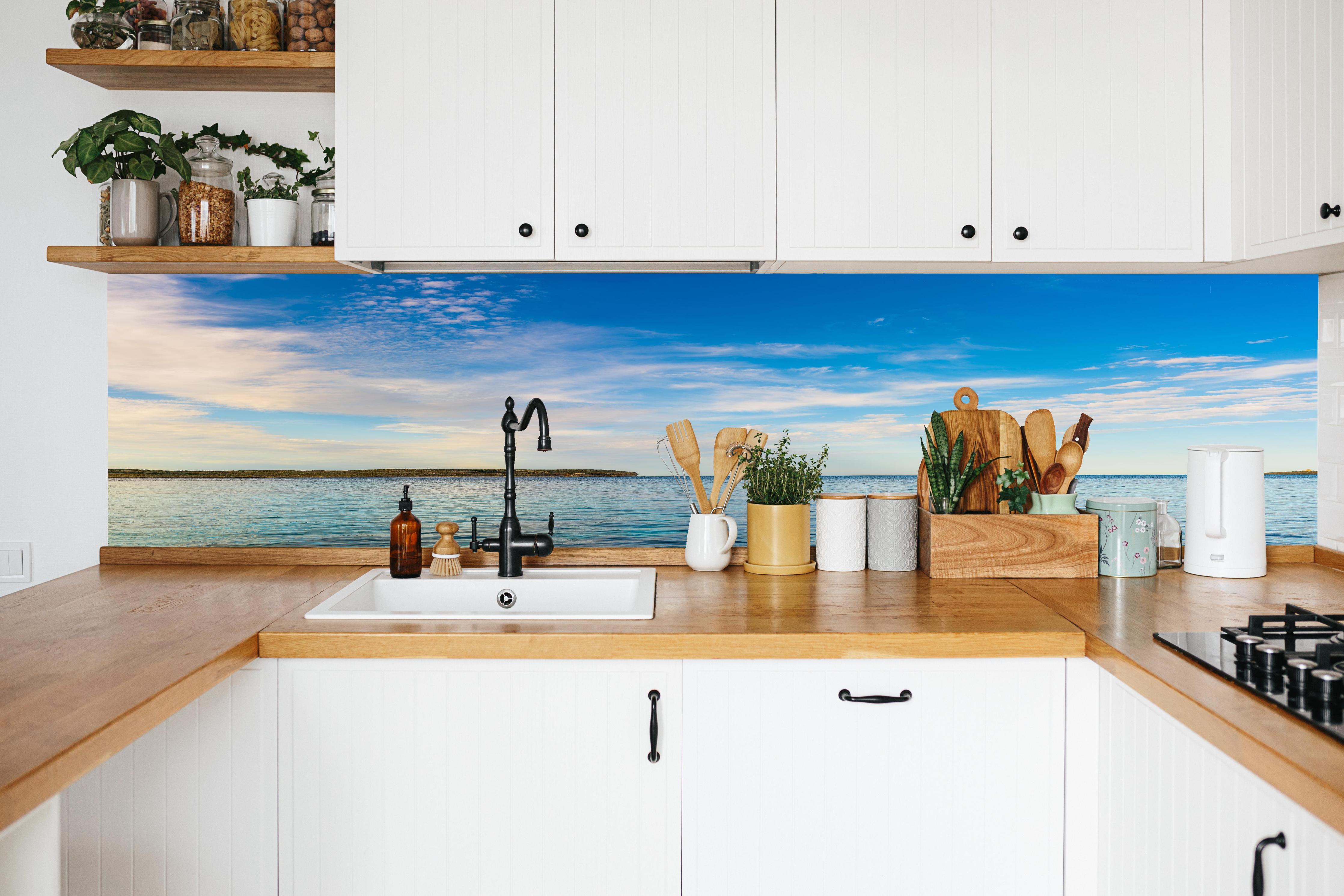 Küche - Weite und ruhige Meerelandschaft unter einem strahlend blauen Himmel hinter weißen Hochglanz-Küchenregalen und schwarzem Wasserhahn