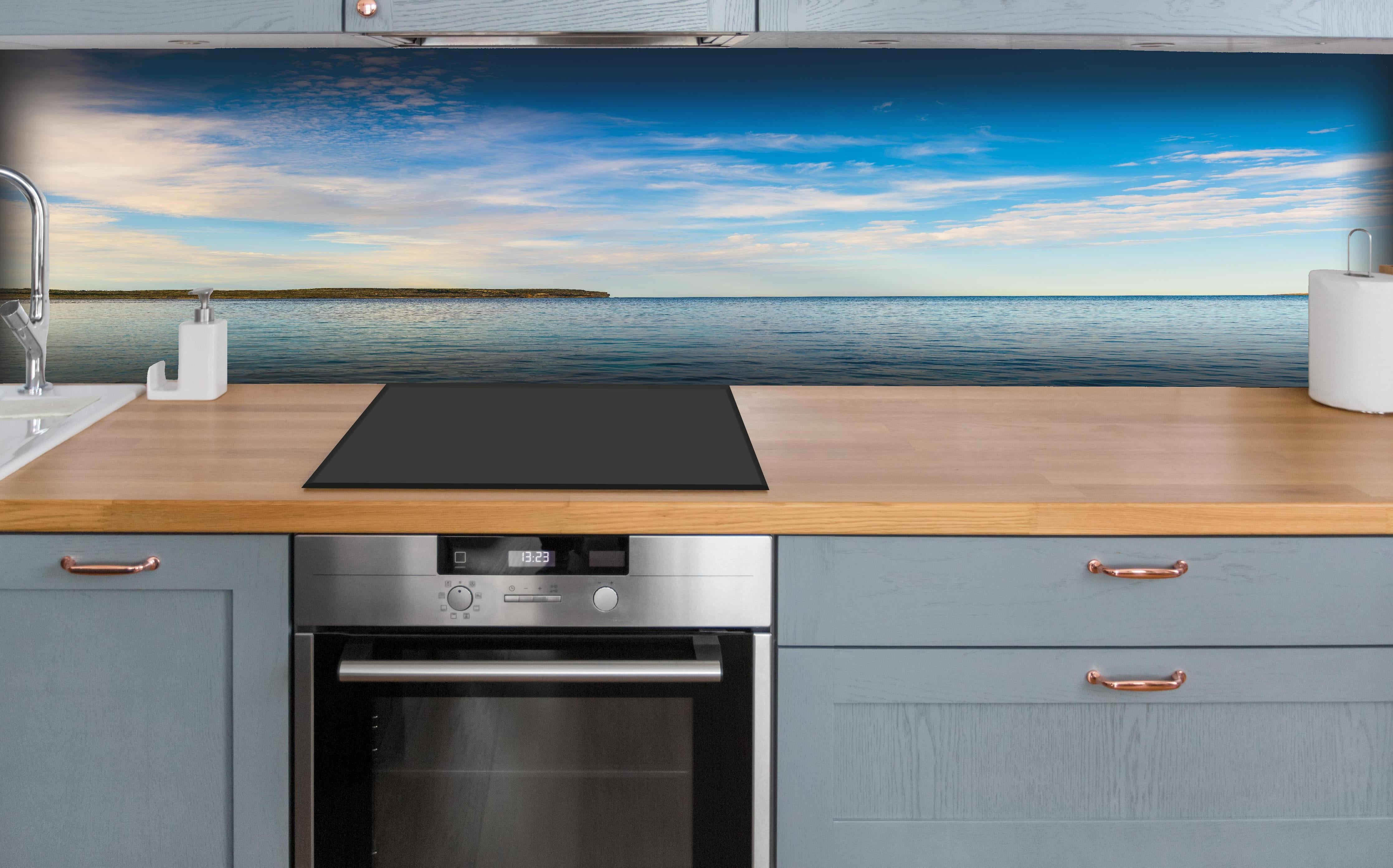Küche - Weite und ruhige Meerelandschaft unter einem strahlend blauen Himmel hinter weißen Hochglanz-Küchenregalen und schwarzem Wasserhahn