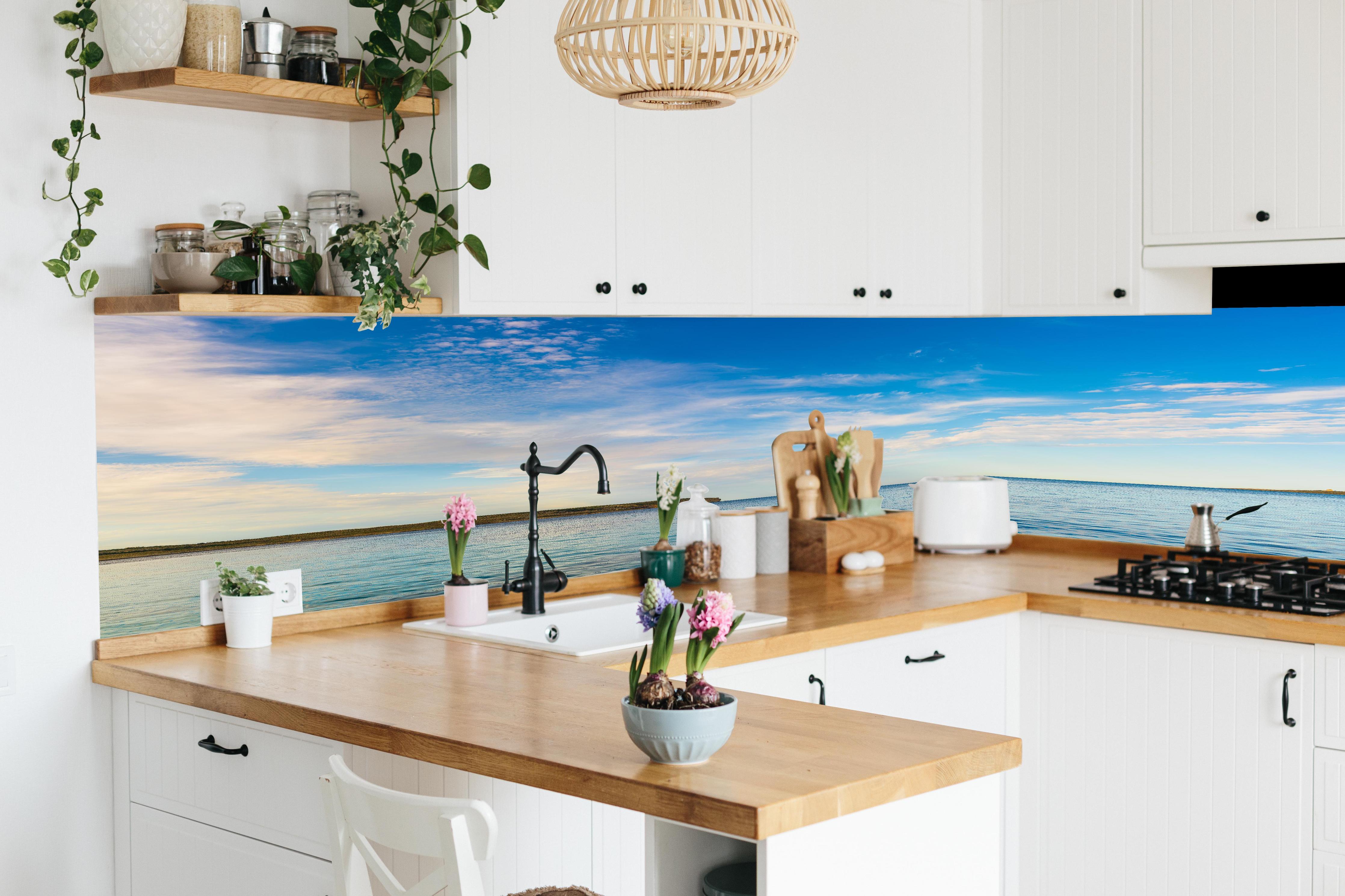 Küche - Weite und ruhige Meerelandschaft unter einem strahlend blauen Himmel hinter weißen Hochglanz-Küchenregalen und schwarzem Wasserhahn