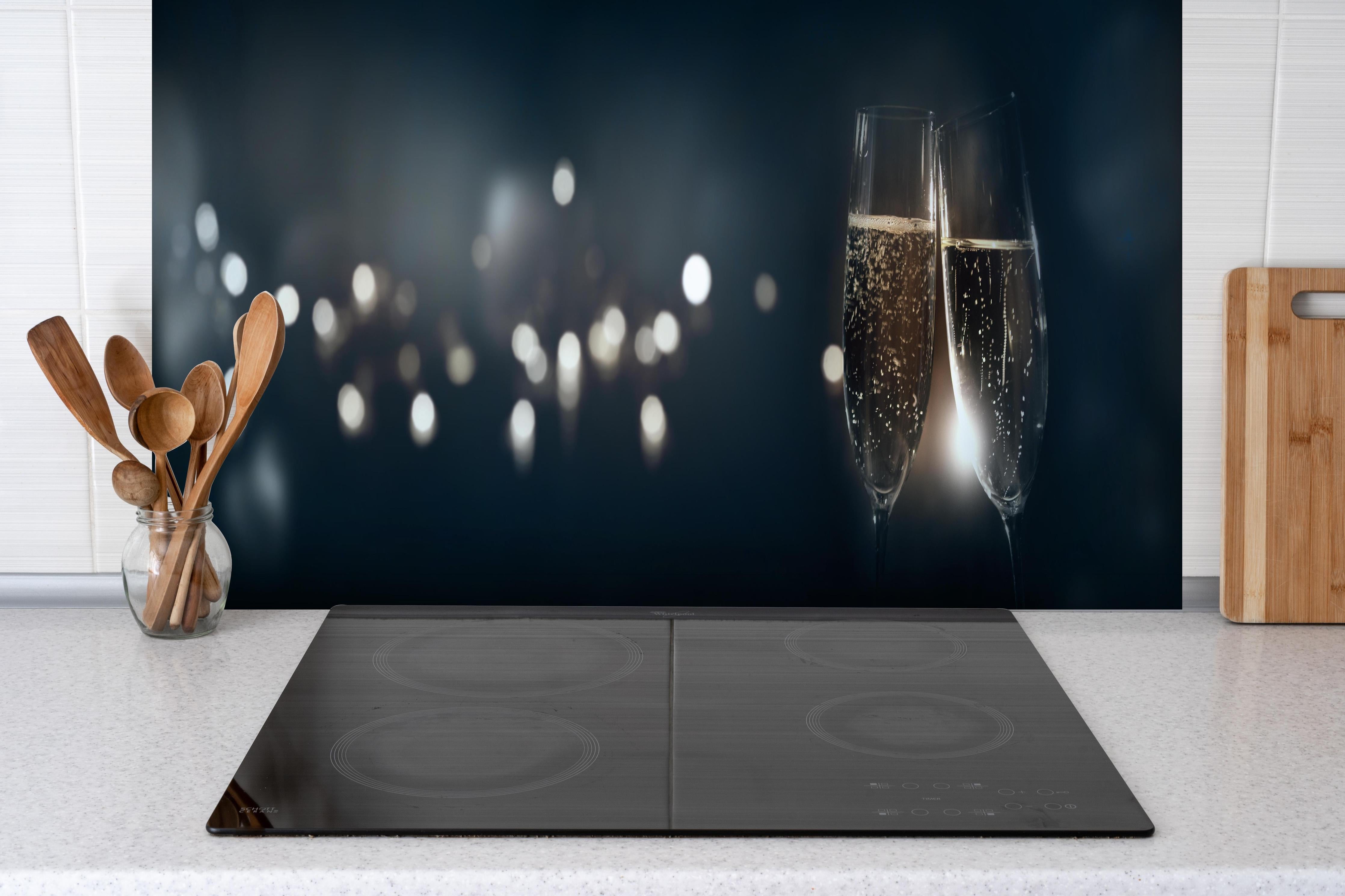 Spritzschutz - Anstoßen mit funkelndem Champagner hinter einem Cerankochfeld zwischen Holz-Kochutensilien
