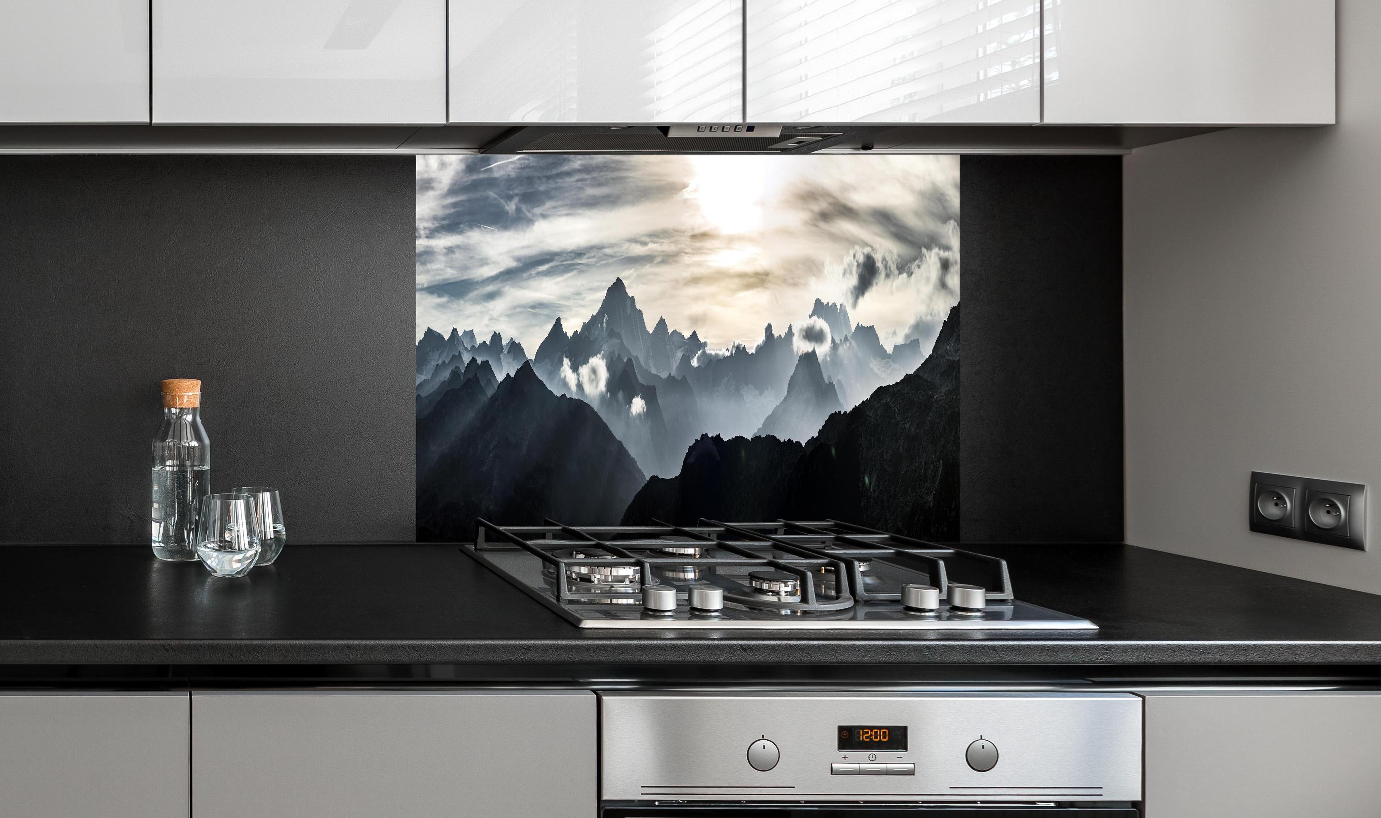 Spritzschutz - Bergpanorama mit Sonnenuntergangslicht hinter einem Cerankochfeld zwischen Holz-Kochutensilien
