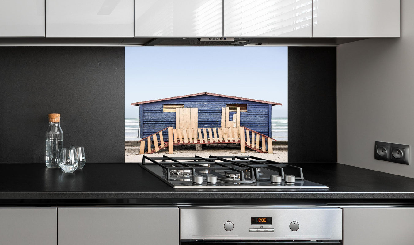 Spritzschutz - Blaue Strandhütte mit Meerblick Sommerhaus hinter einem Cerankochfeld zwischen Holz-Kochutensilien
