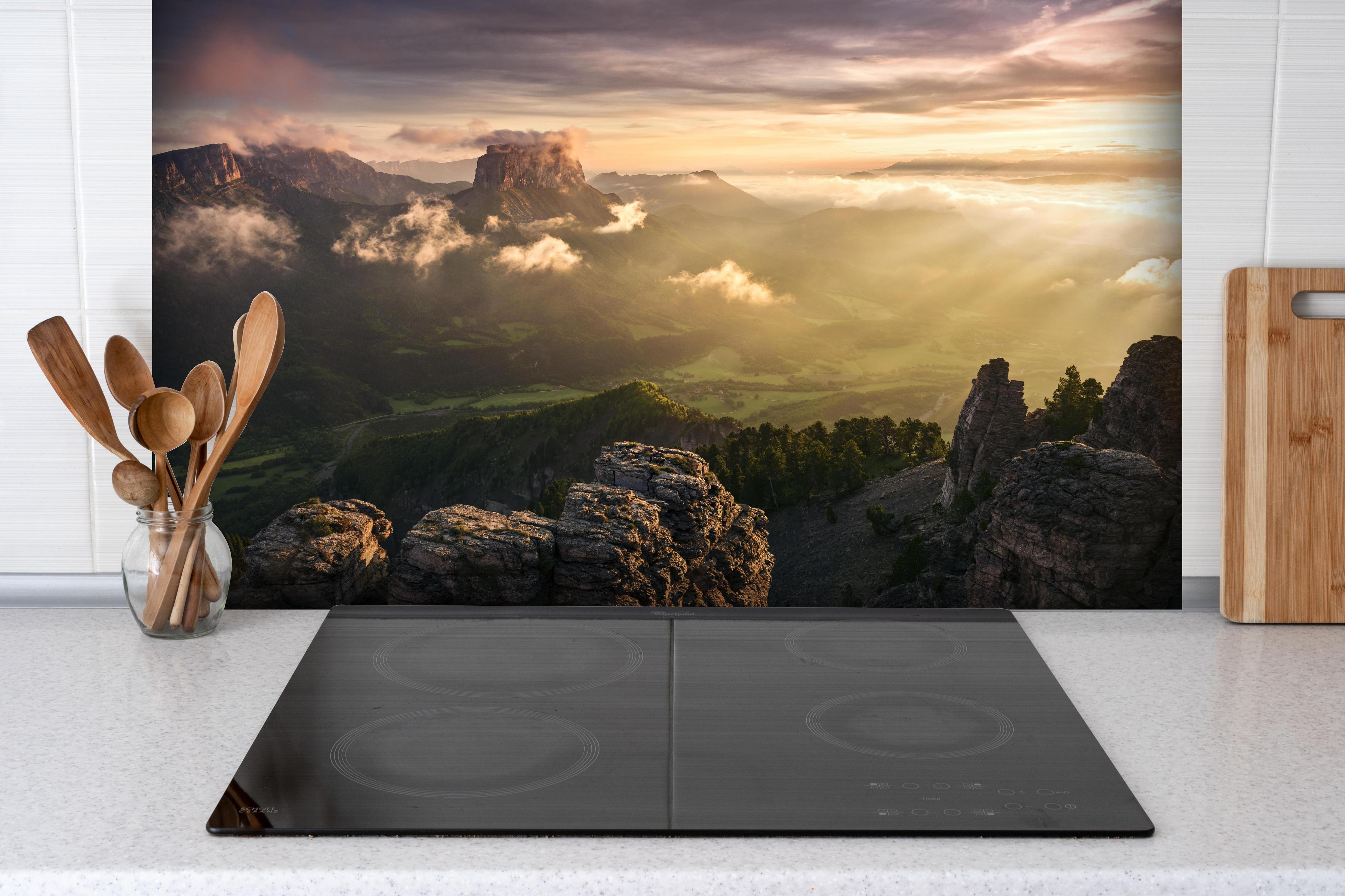 Spritzschutz - Dramatische Morgenlichtstrahlen in Alpen hinter einem Cerankochfeld zwischen Holz-Kochutensilien
