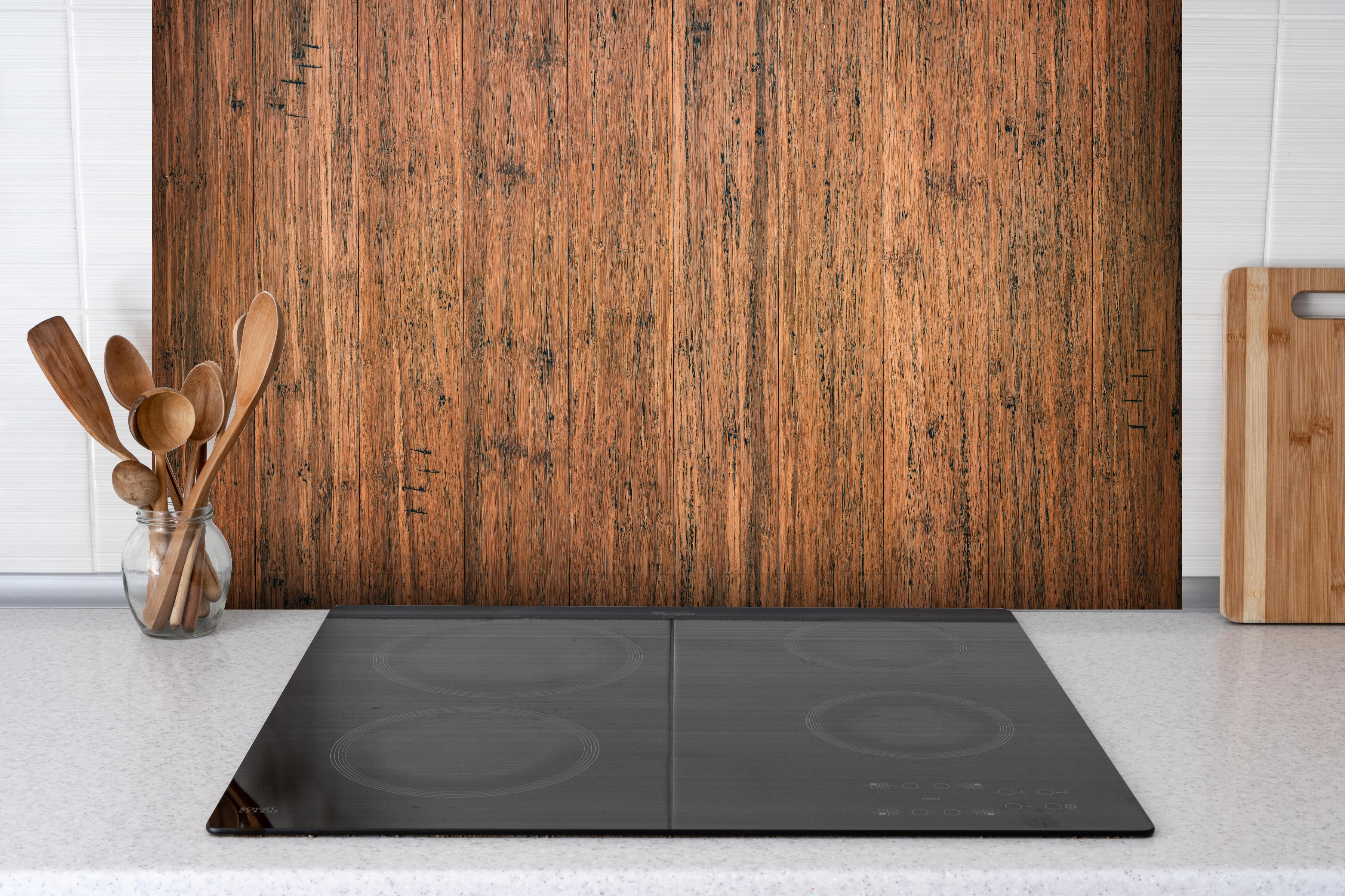 Spritzschutz - Dunkelbraunes rustikales Holzbrett hinter einem Cerankochfeld zwischen Holz-Kochutensilien

