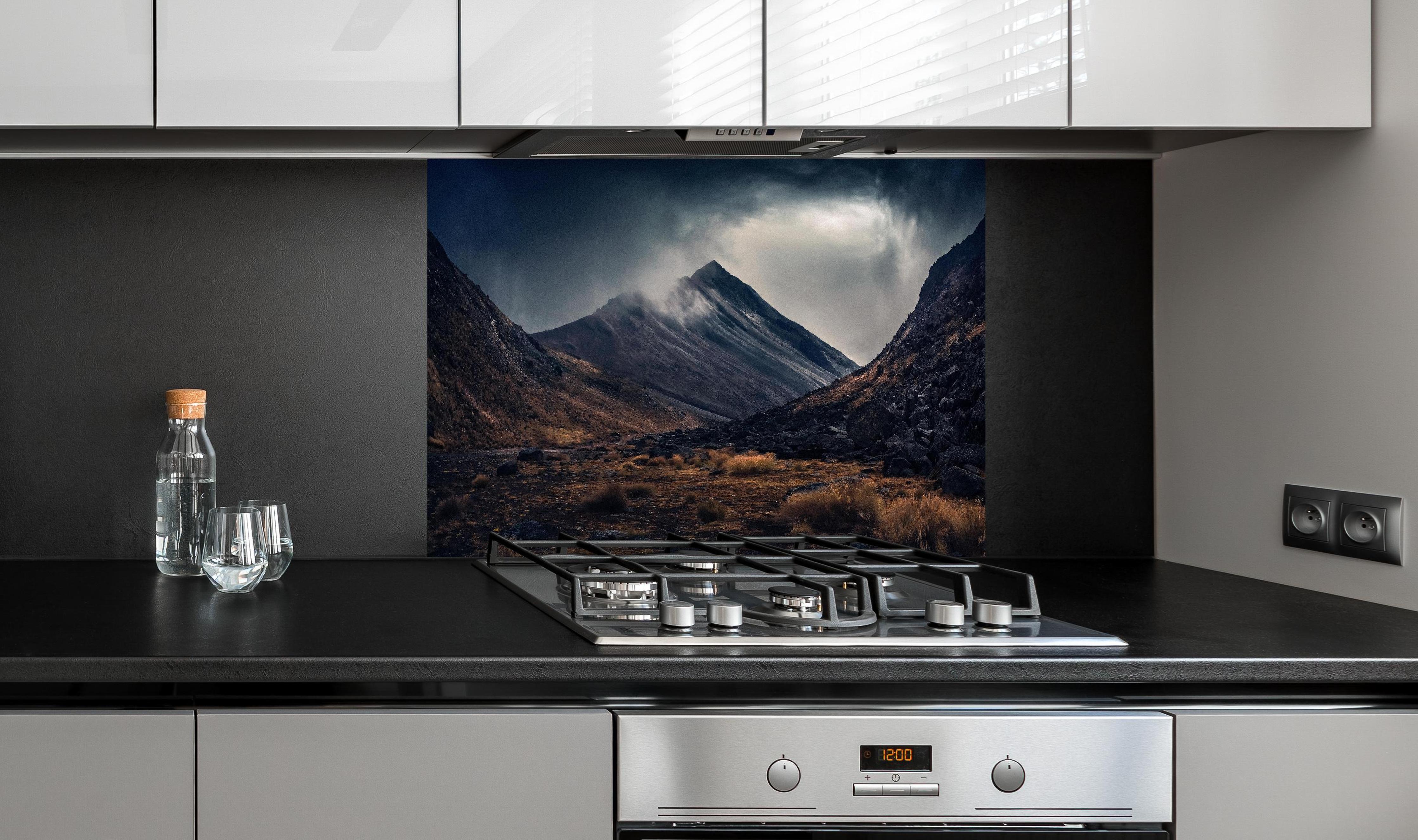 Spritzschutz - Dunkle Wolken Aufziehend über Bergspitze hinter einem Cerankochfeld zwischen Holz-Kochutensilien
