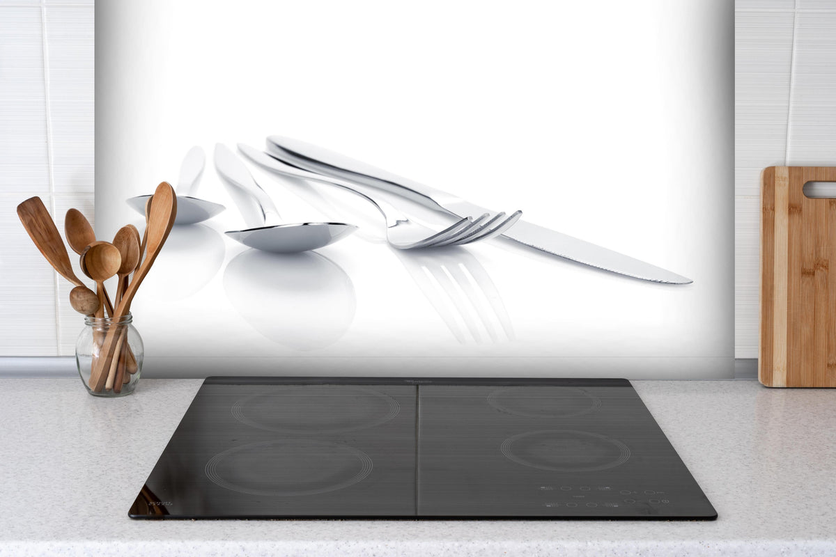 Spritzschutz - Elegantes Besteck-Set in Weiß mit Silberakzenten hinter einem Cerankochfeld zwischen Holz-Kochutensilien
