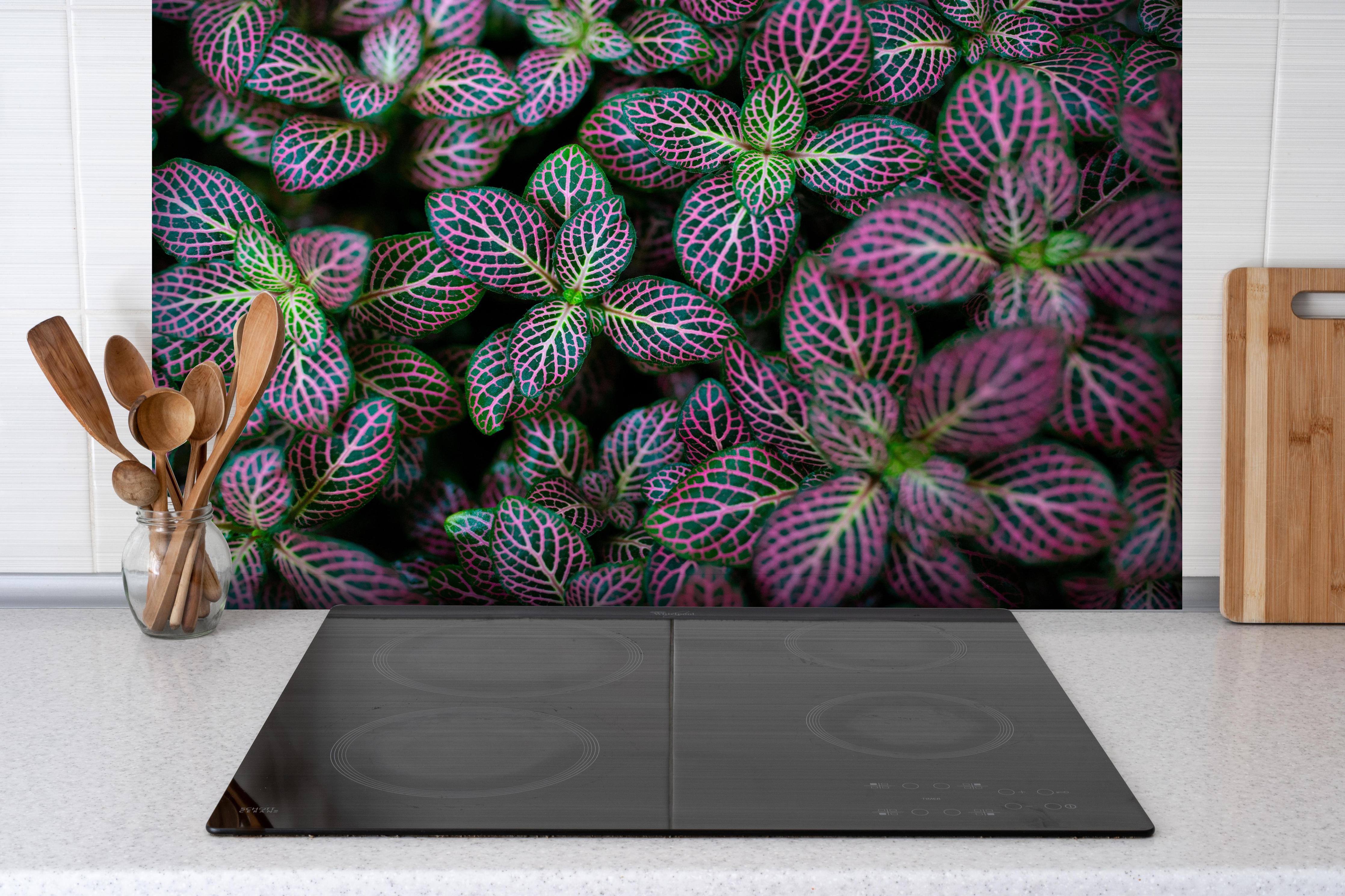 Spritzschutz - Exotische Blätter Textur in Bunt hinter einem Cerankochfeld zwischen Holz-Kochutensilien
