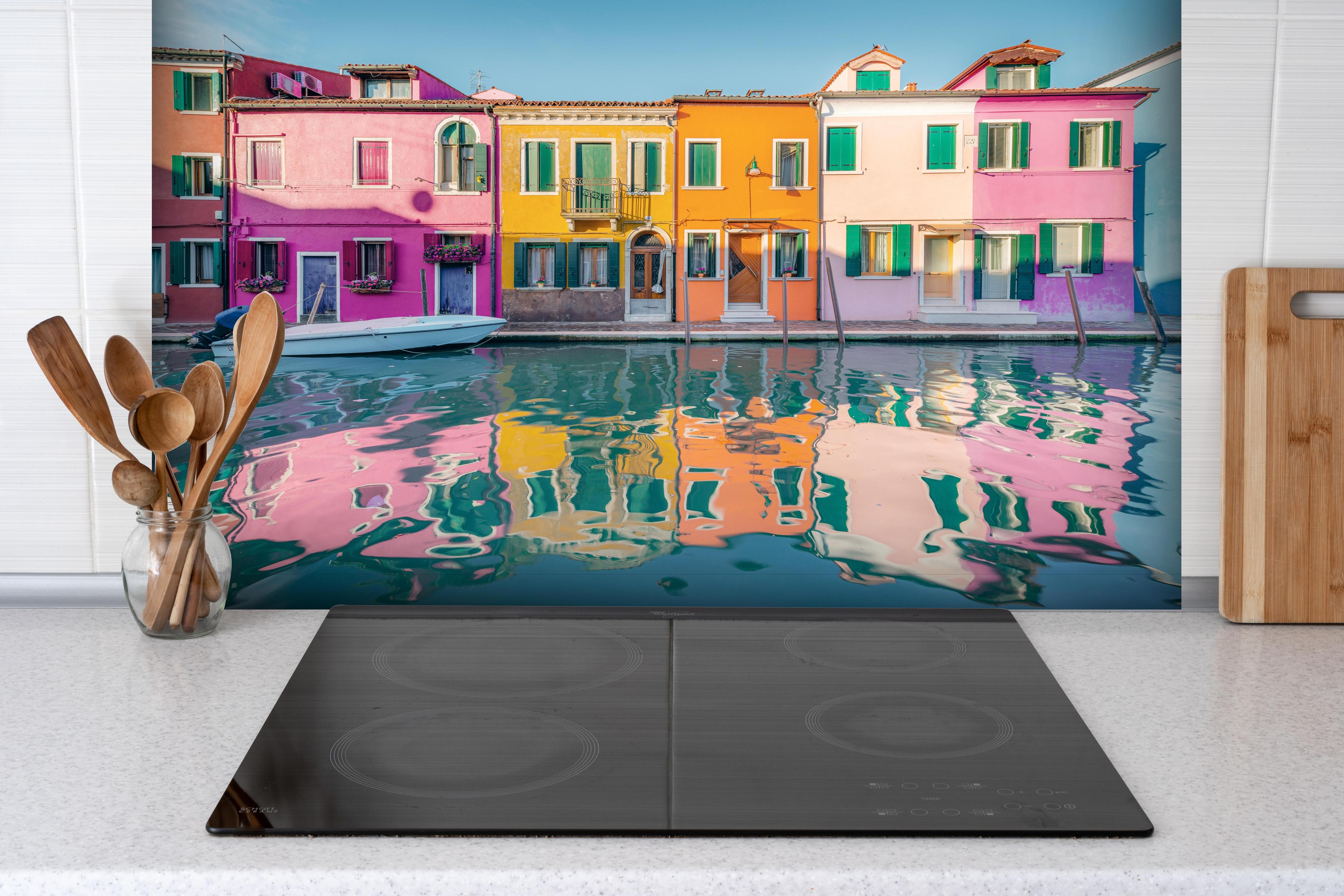 Spritzschutz - Farbenfrohe Häuser am Wasser in Burano, Venedig hinter einem Cerankochfeld zwischen Holz-Kochutensilien
