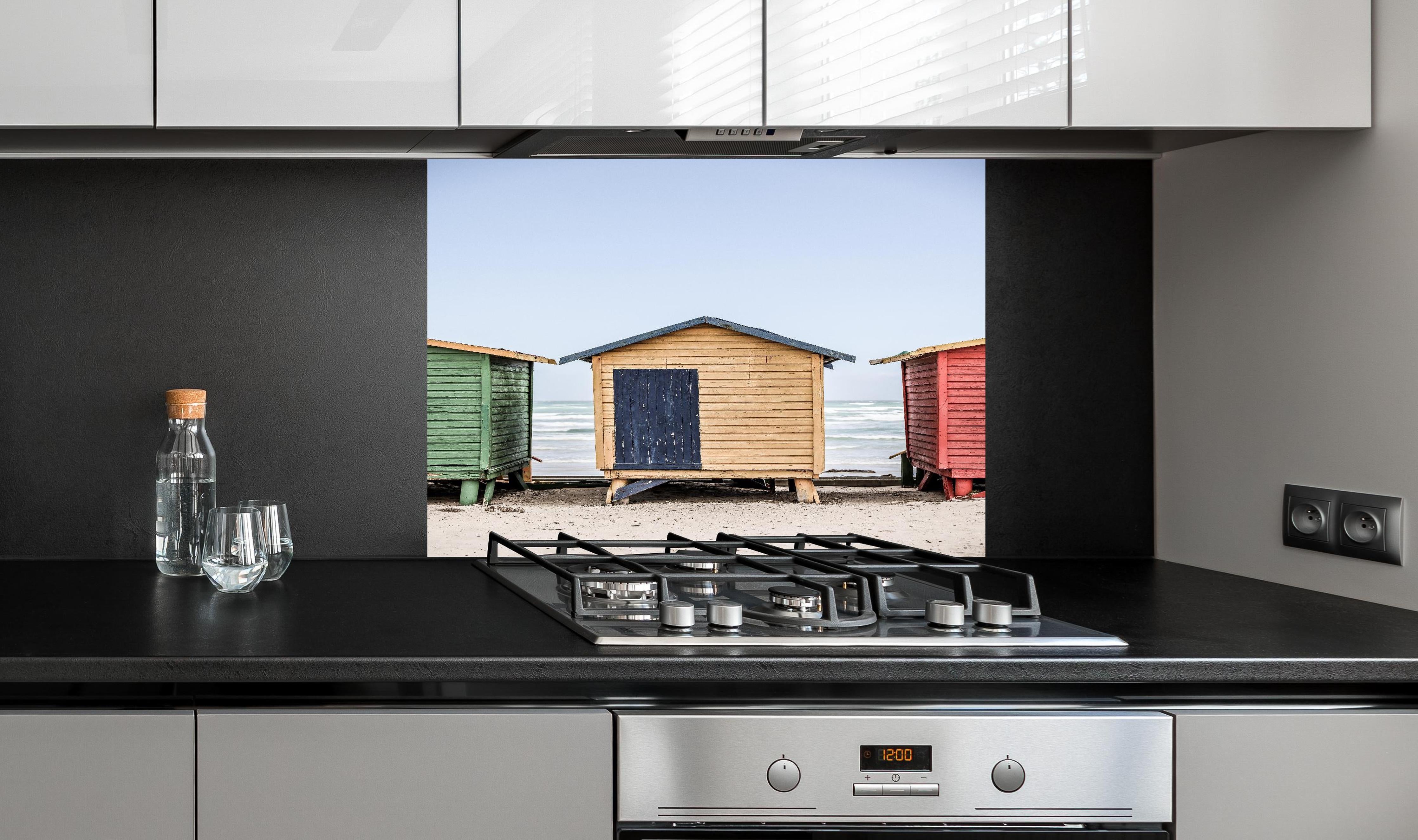 Spritzschutz - Farbenfrohe Strandhütten am Meeressand hinter einem Cerankochfeld zwischen Holz-Kochutensilien
