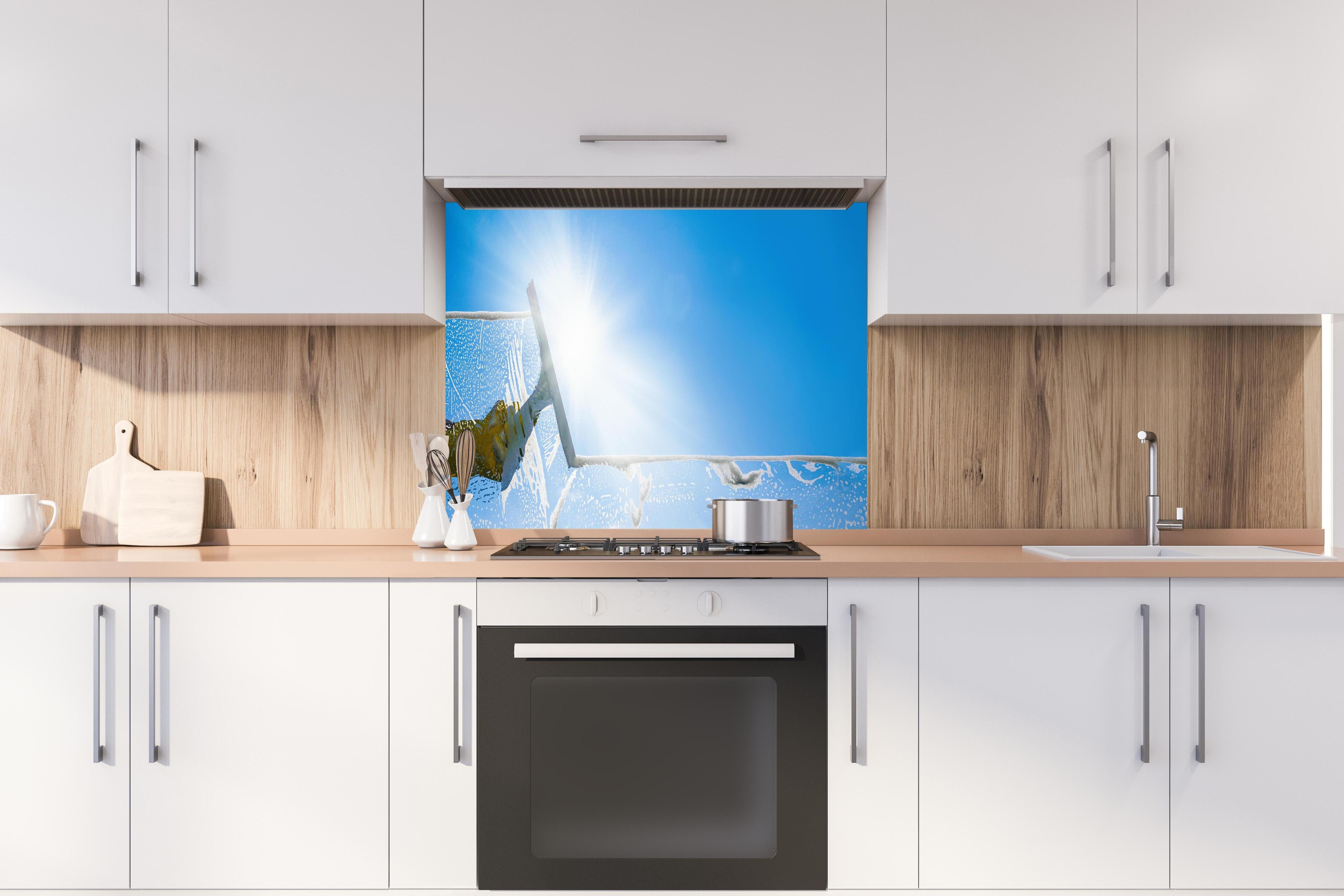 Spritzschutz - Fensterputzer mit Abzieher und strahlendem Himmel hinter einem Cerankochfeld zwischen Holz-Kochutensilien
