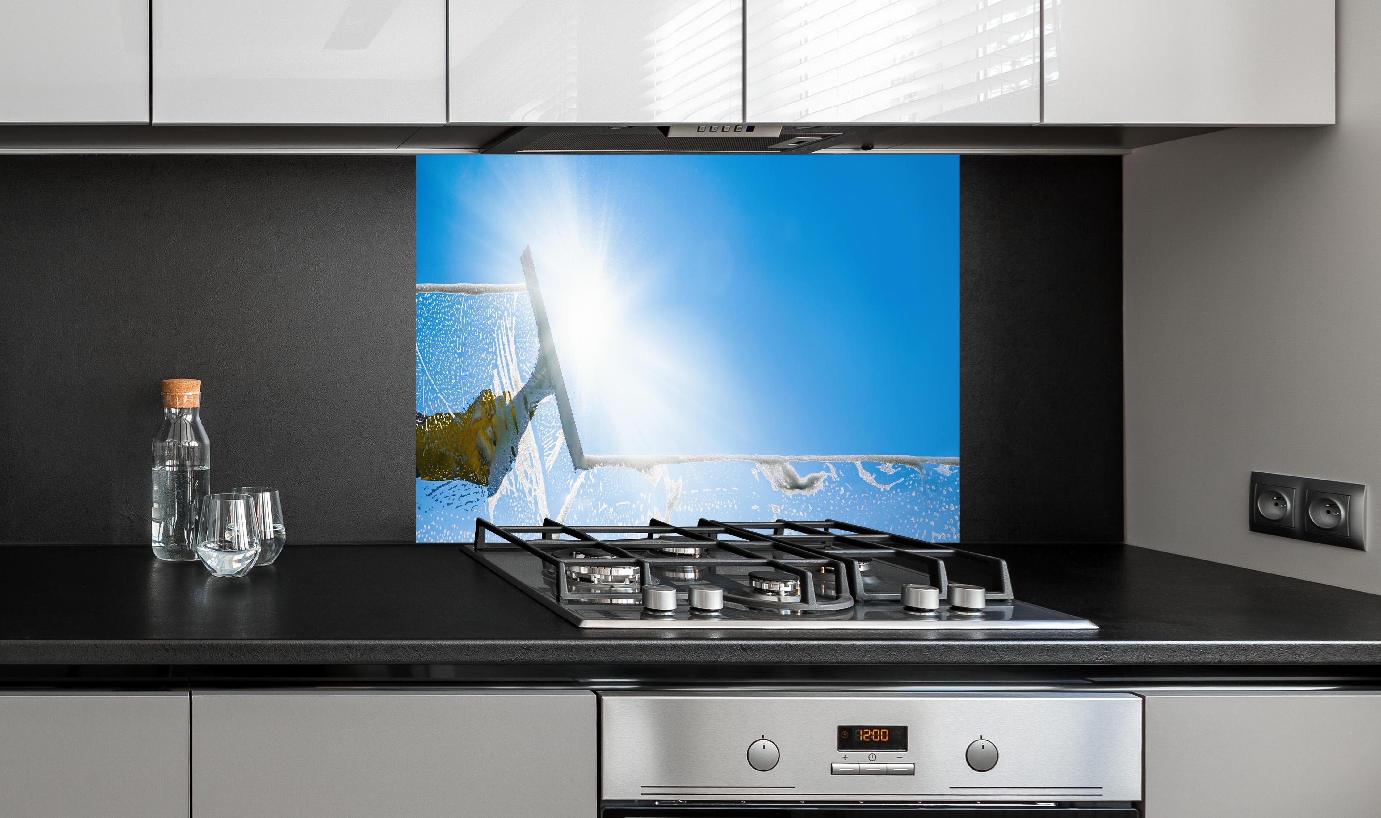 Spritzschutz - Fensterputzer mit Abzieher und strahlendem Himmel hinter einem Cerankochfeld zwischen Holz-Kochutensilien
