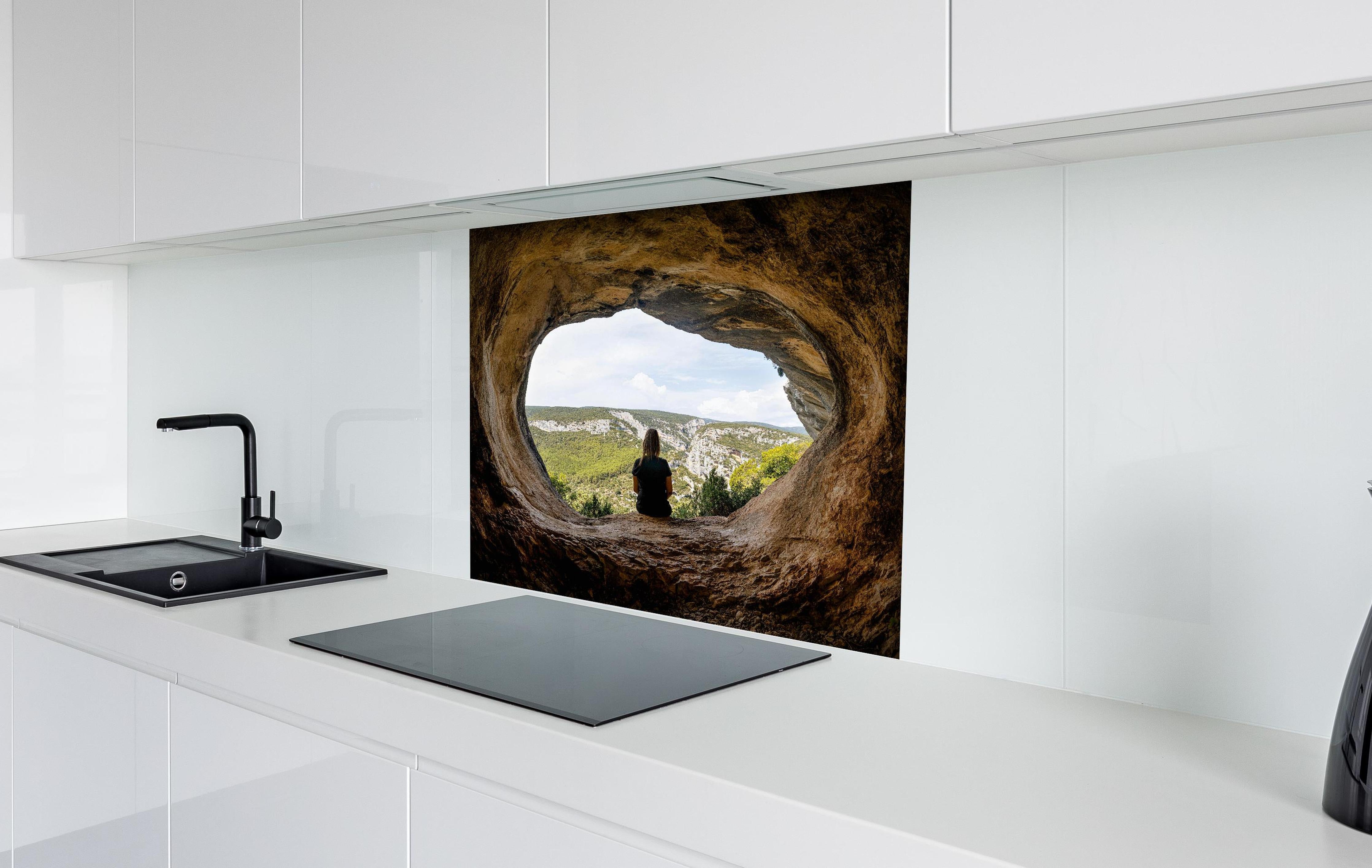 Spritzschutz - Frau sitzend in Berghöhle mit Blick auf Landschaft hinter einem Cerankochfeld zwischen Holz-Kochutensilien
