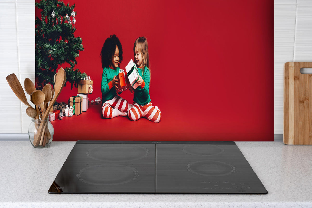 Spritzschutz - Fröhliche Kinder tauschen Weihnachtsgeschenke aus hinter einem Cerankochfeld zwischen Holz-Kochutensilien
