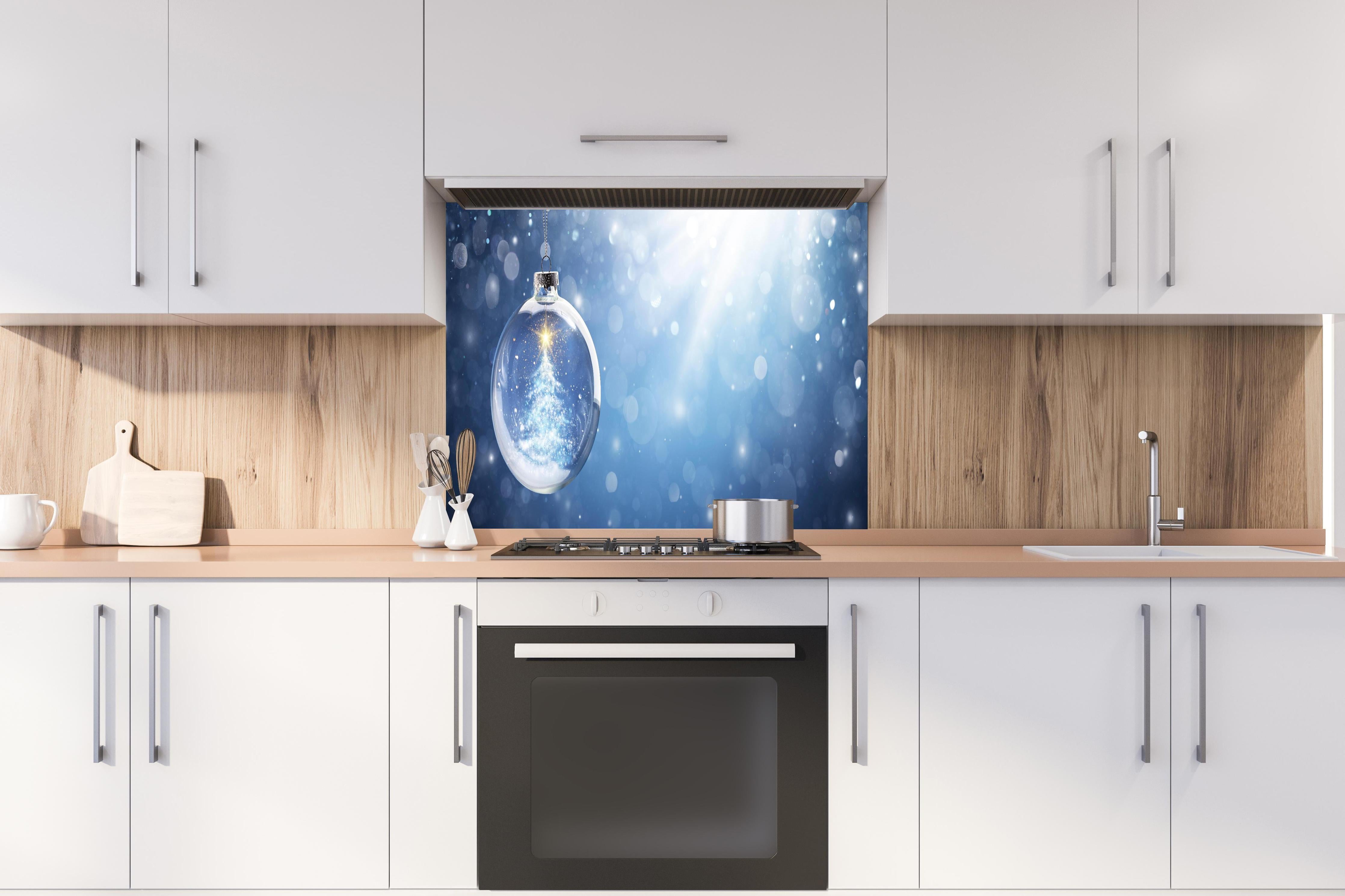 Spritzschutz - Glänzender Weihnachtsbaum in Schneekugel hinter einem Cerankochfeld zwischen Holz-Kochutensilien
