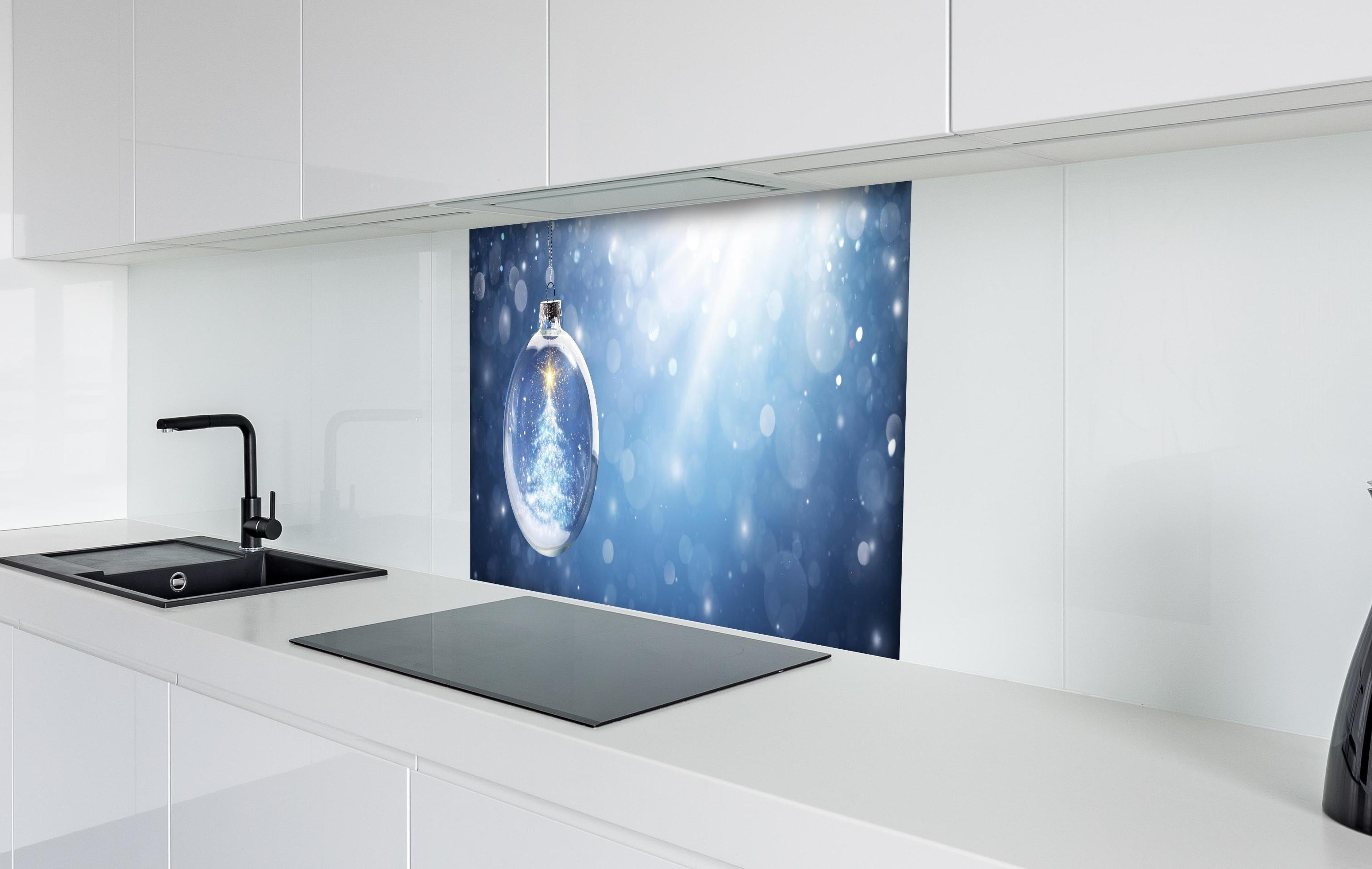 Spritzschutz - Glänzender Weihnachtsbaum in Schneekugel hinter einem Cerankochfeld zwischen Holz-Kochutensilien
