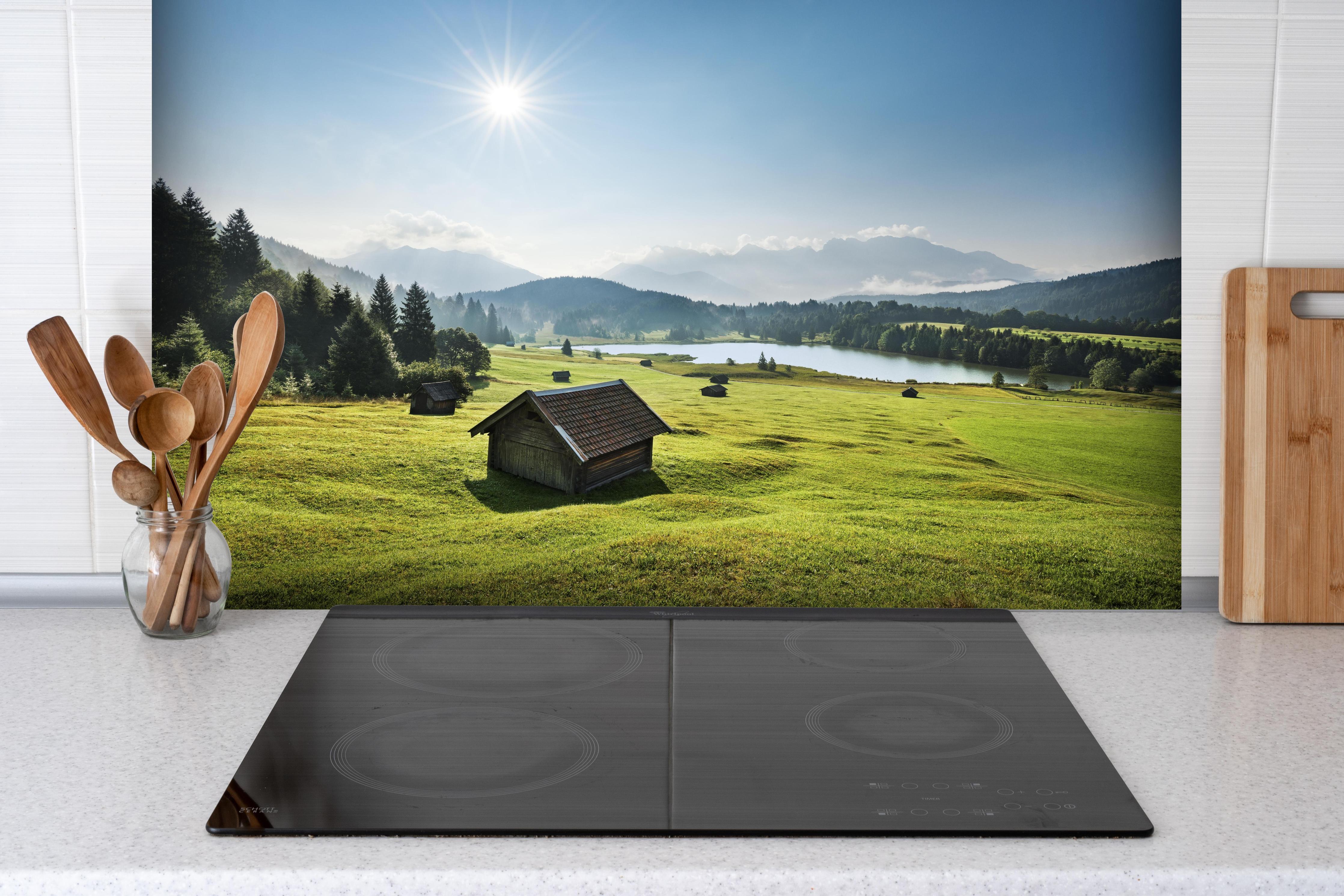 Spritzschutz - Grüne Alpenwiese, Berge und See im Sonnenschein hinter einem Cerankochfeld zwischen Holz-Kochutensilien
