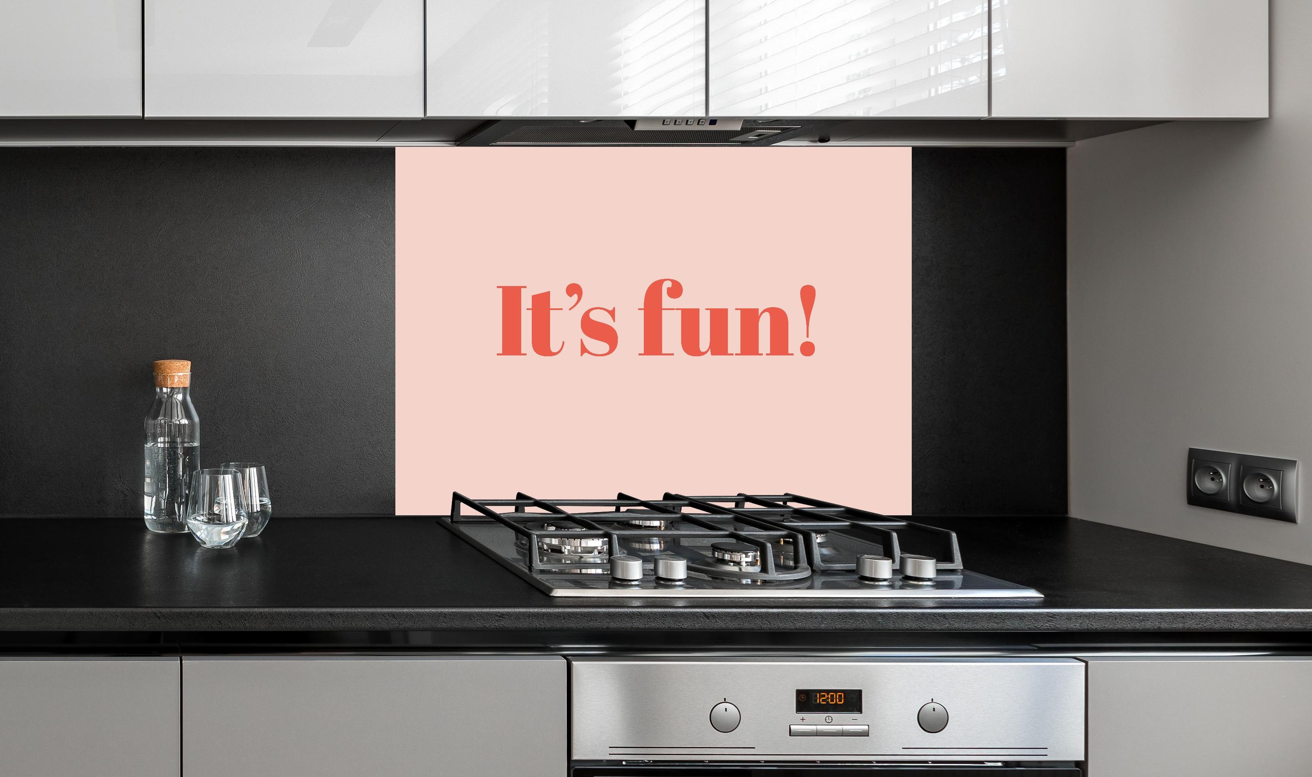 Spritzschutz - Inspirierender 'It's Fun!' Typo-Print in Pink hinter einem Cerankochfeld zwischen Holz-Kochutensilien
