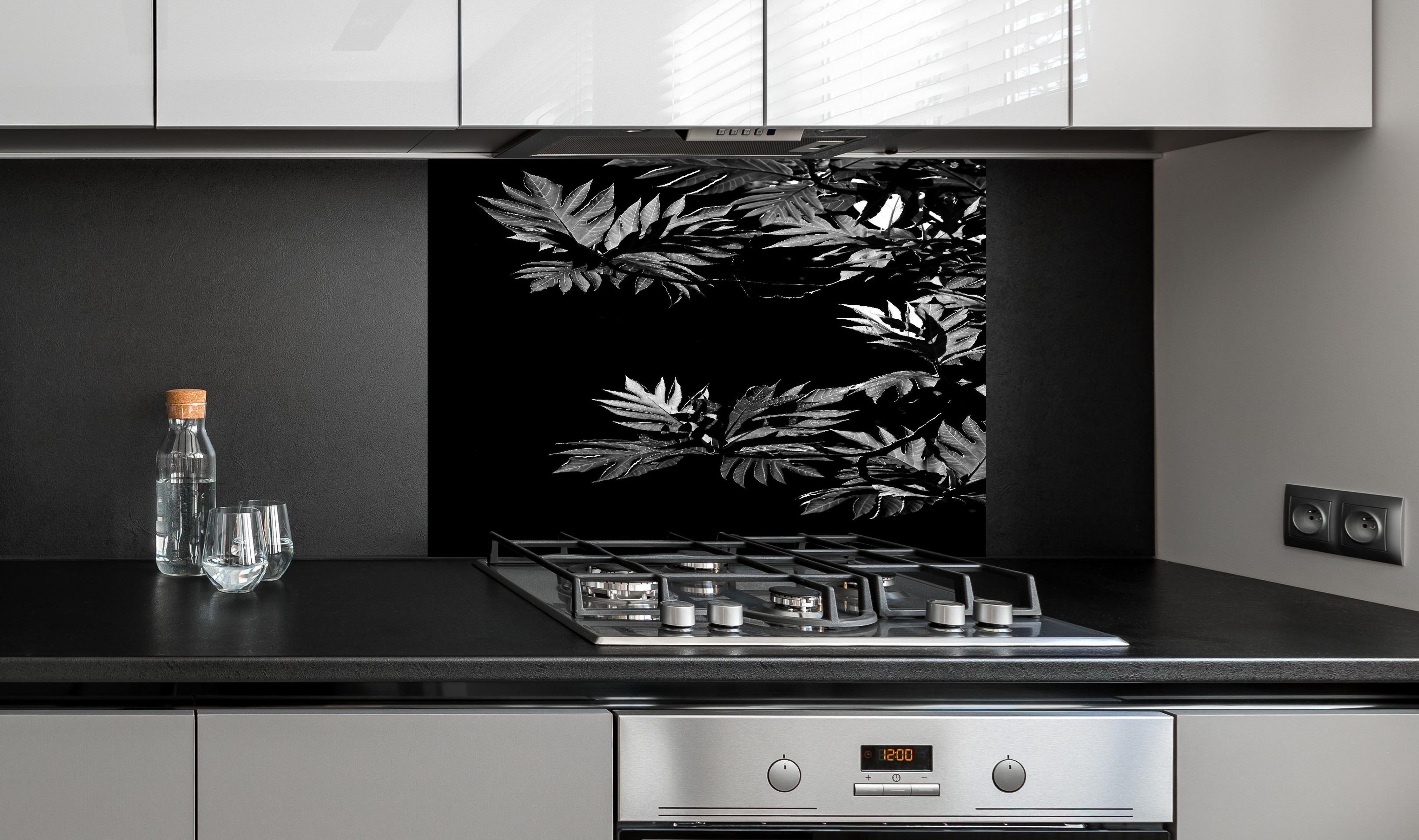 Spritzschutz - Kontrastreiche Blätter auf schwarzem Hintergrund hinter einem Cerankochfeld zwischen Holz-Kochutensilien
