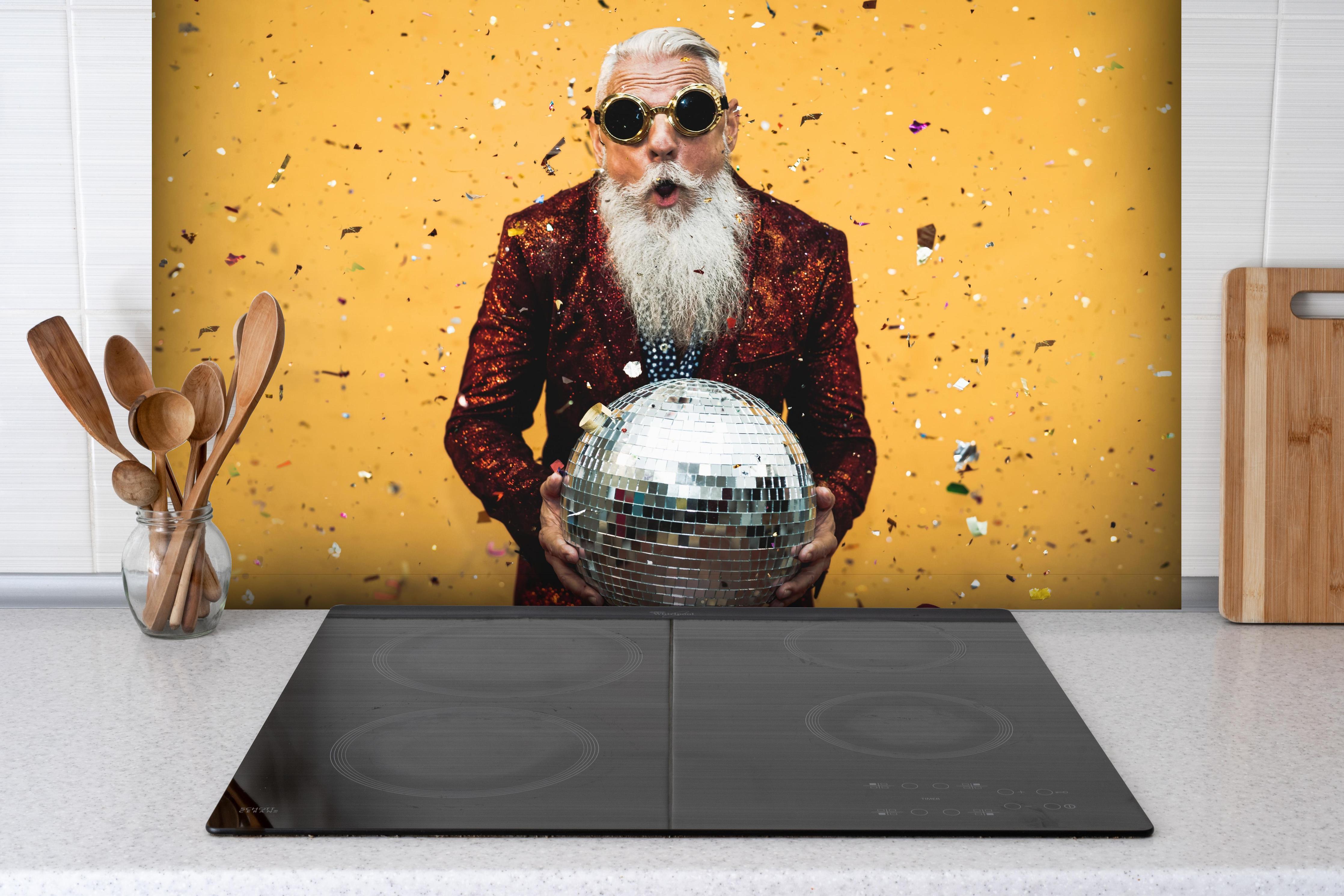 Spritzschutz - Lebensfroher Senior feiert mit Discokugel hinter einem Cerankochfeld zwischen Holz-Kochutensilien

