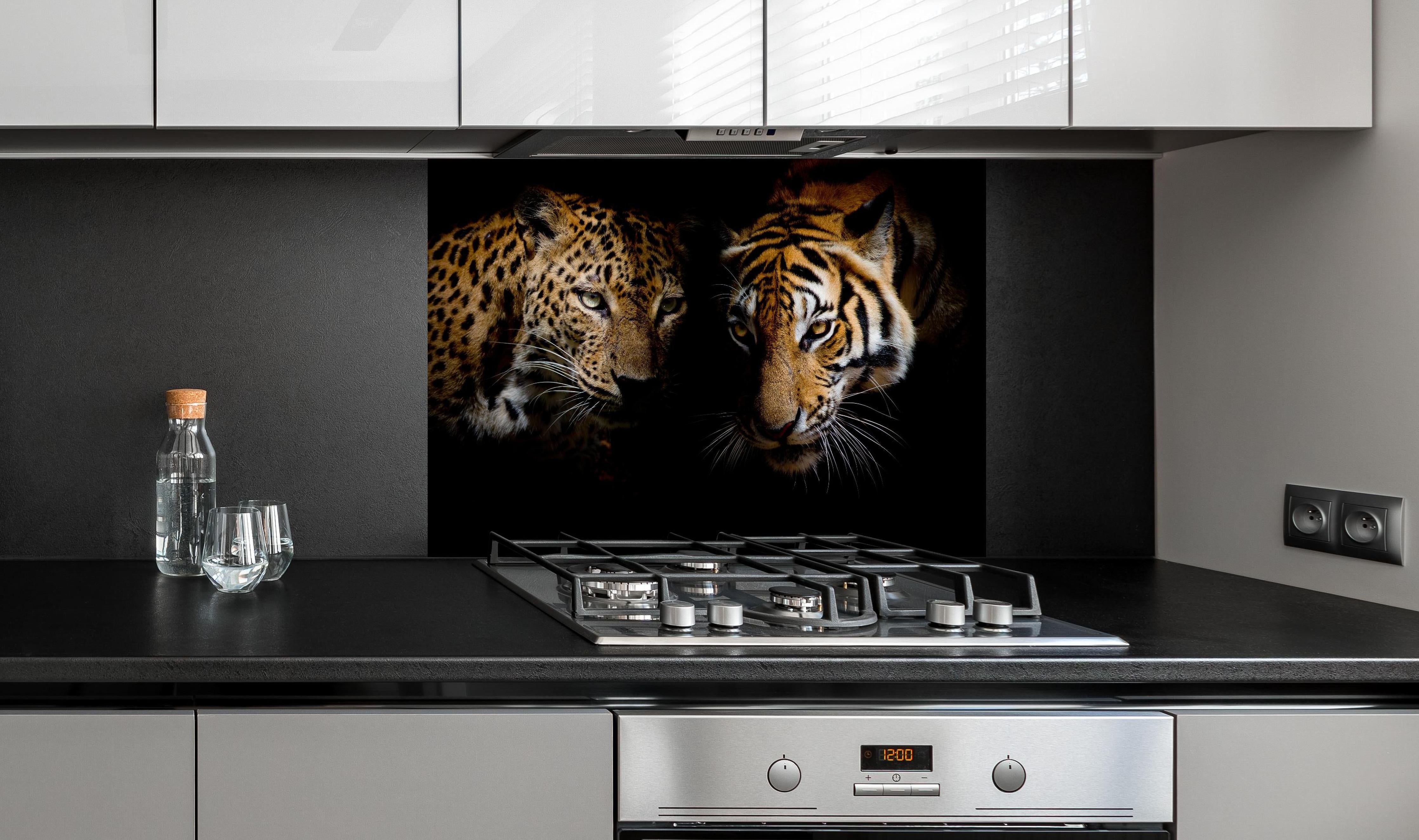Spritzschutz - Leopard und Tiger isoliert auf schwarzen Hintergrund hinter einem Cerankochfeld zwischen Holz-Kochutensilien
