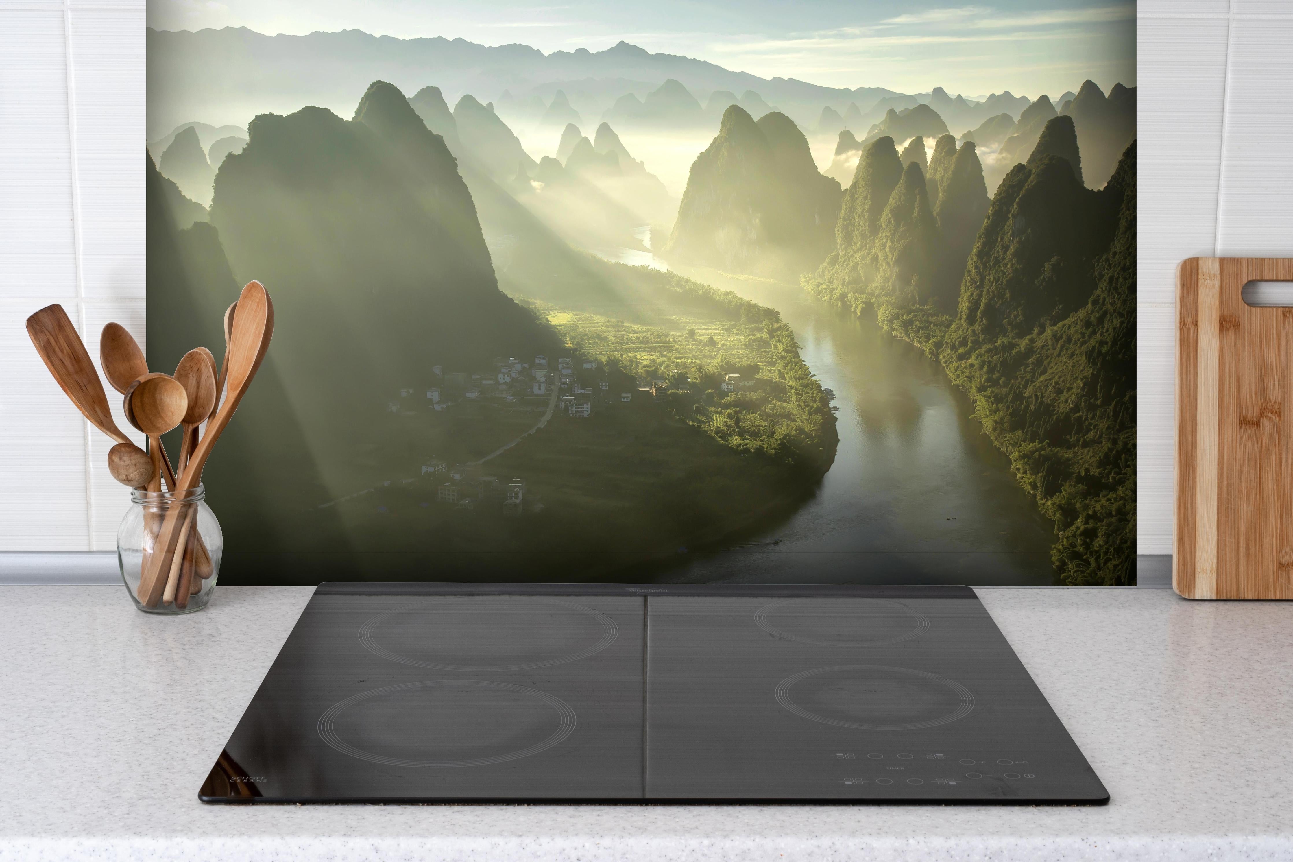 Spritzschutz - Lichtstrahlen über Li-Fluss und Karstberge hinter einem Cerankochfeld zwischen Holz-Kochutensilien
