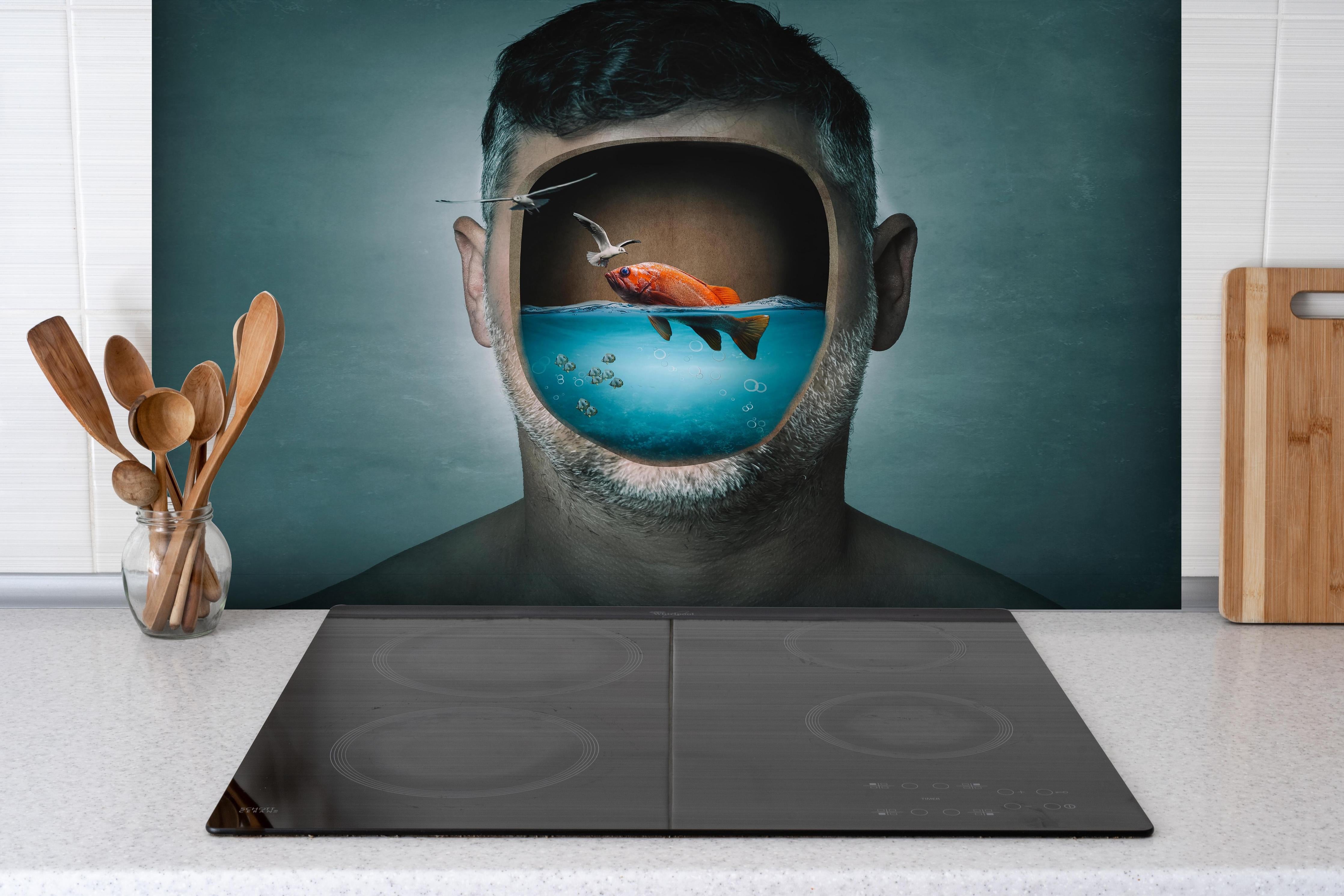 Spritzschutz - Mann mit Fisch im Wasser-Gesicht hinter einem Cerankochfeld zwischen Holz-Kochutensilien

