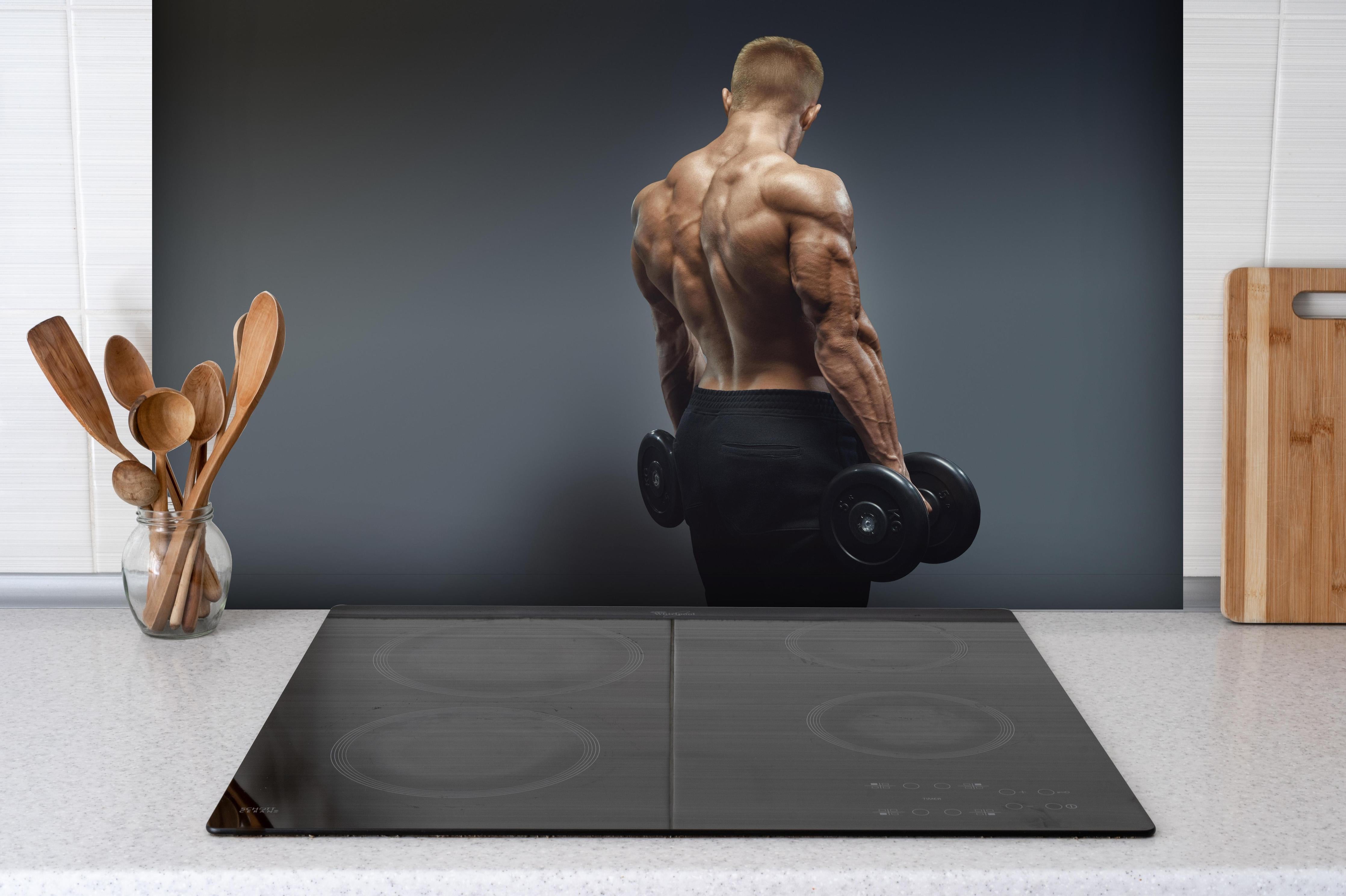 Spritzschutz - Muskulöser Sportler mit Hanteln hinter einem Cerankochfeld zwischen Holz-Kochutensilien
