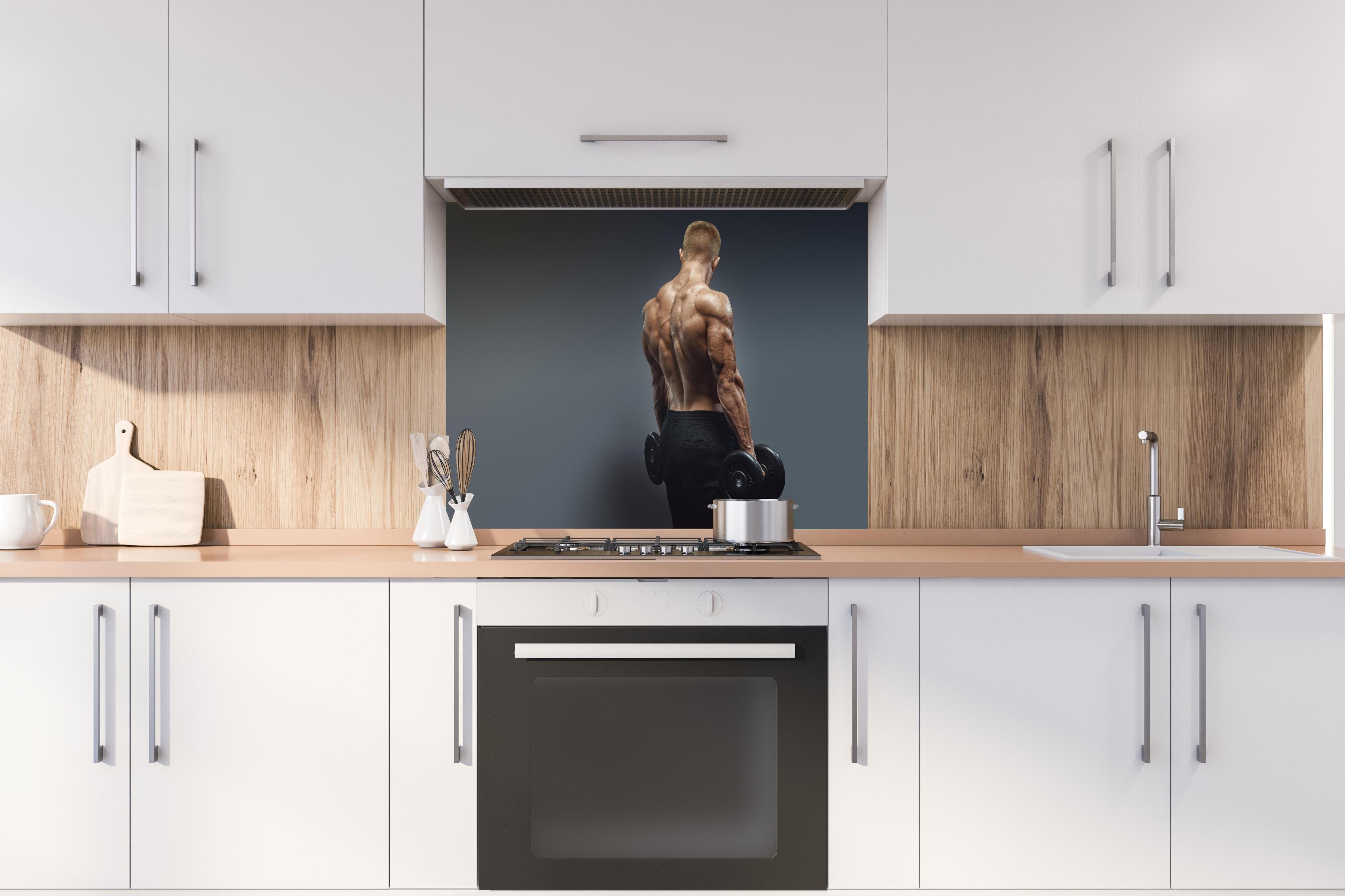 Spritzschutz - Muskulöser Sportler mit Hanteln hinter einem Cerankochfeld zwischen Holz-Kochutensilien
