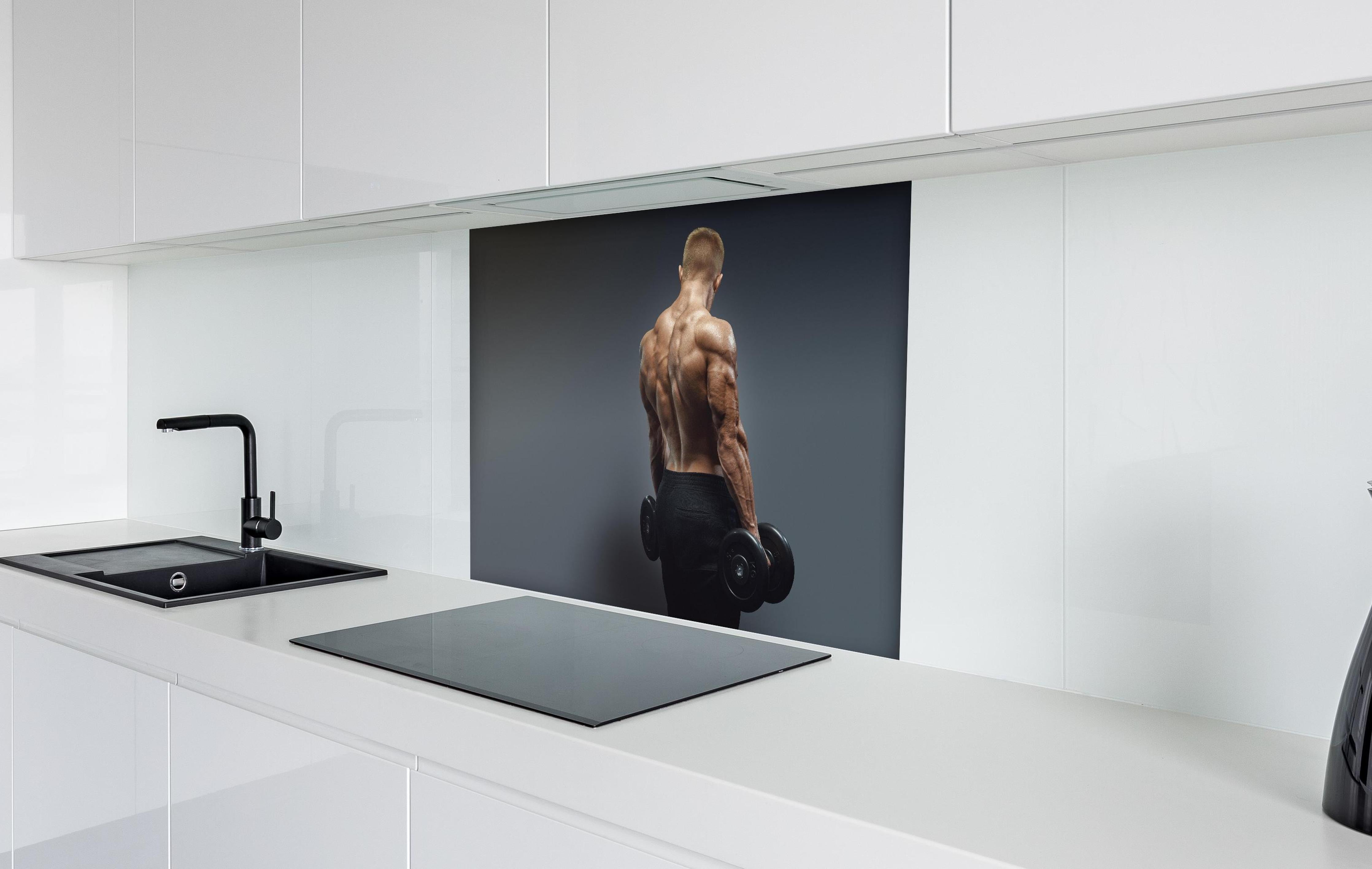 Spritzschutz - Muskulöser Sportler mit Hanteln hinter einem Cerankochfeld zwischen Holz-Kochutensilien
