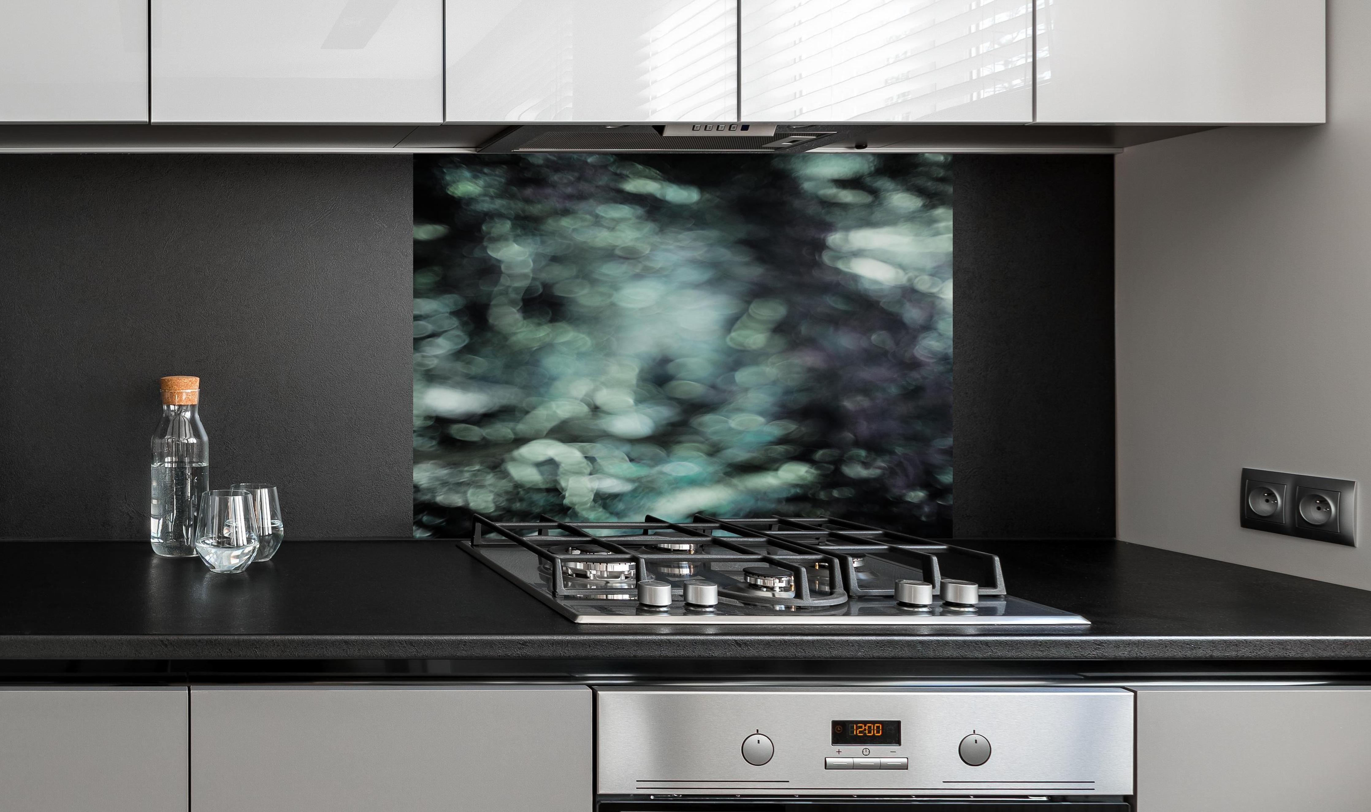 Spritzschutz - Mystischer Grünschimmer auf dunklem Grund hinter einem Cerankochfeld zwischen Holz-Kochutensilien
