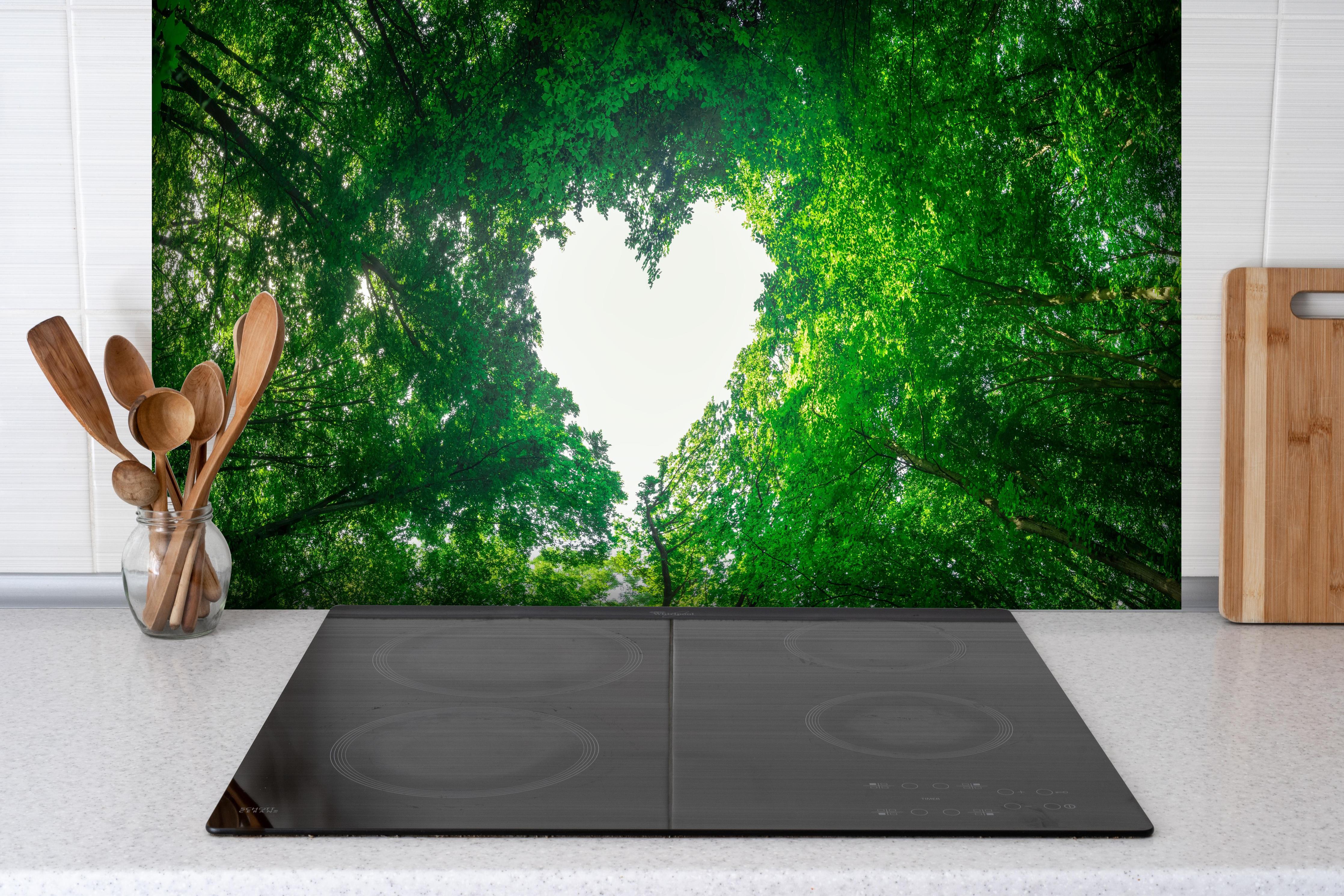 Spritzschutz - Panorama - Baumkronen formen ein Herz hinter einem Cerankochfeld zwischen Holz-Kochutensilien
