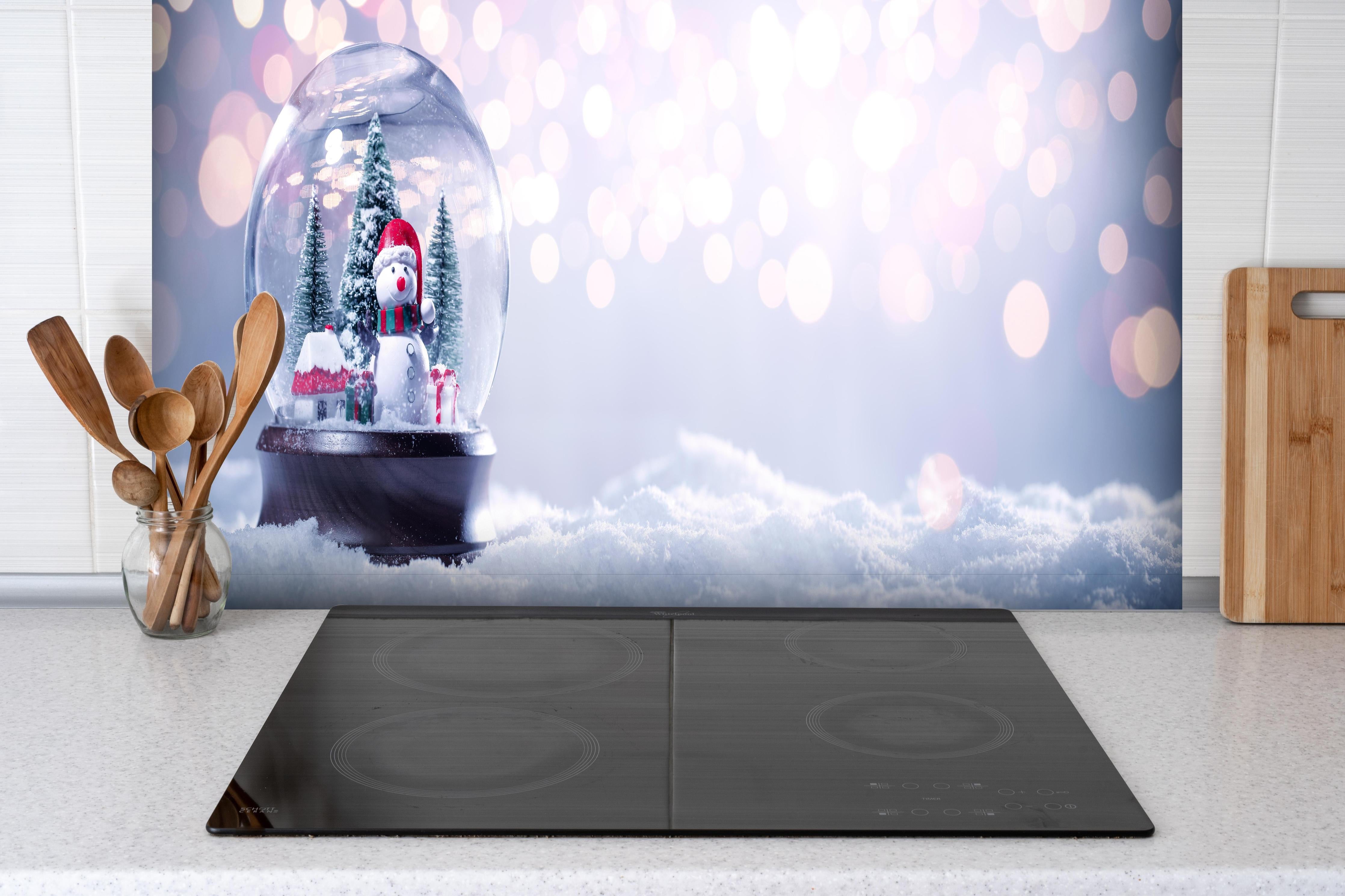 Spritzschutz - Schneekugel auf festlichem Hintergrund hinter einem Cerankochfeld zwischen Holz-Kochutensilien
