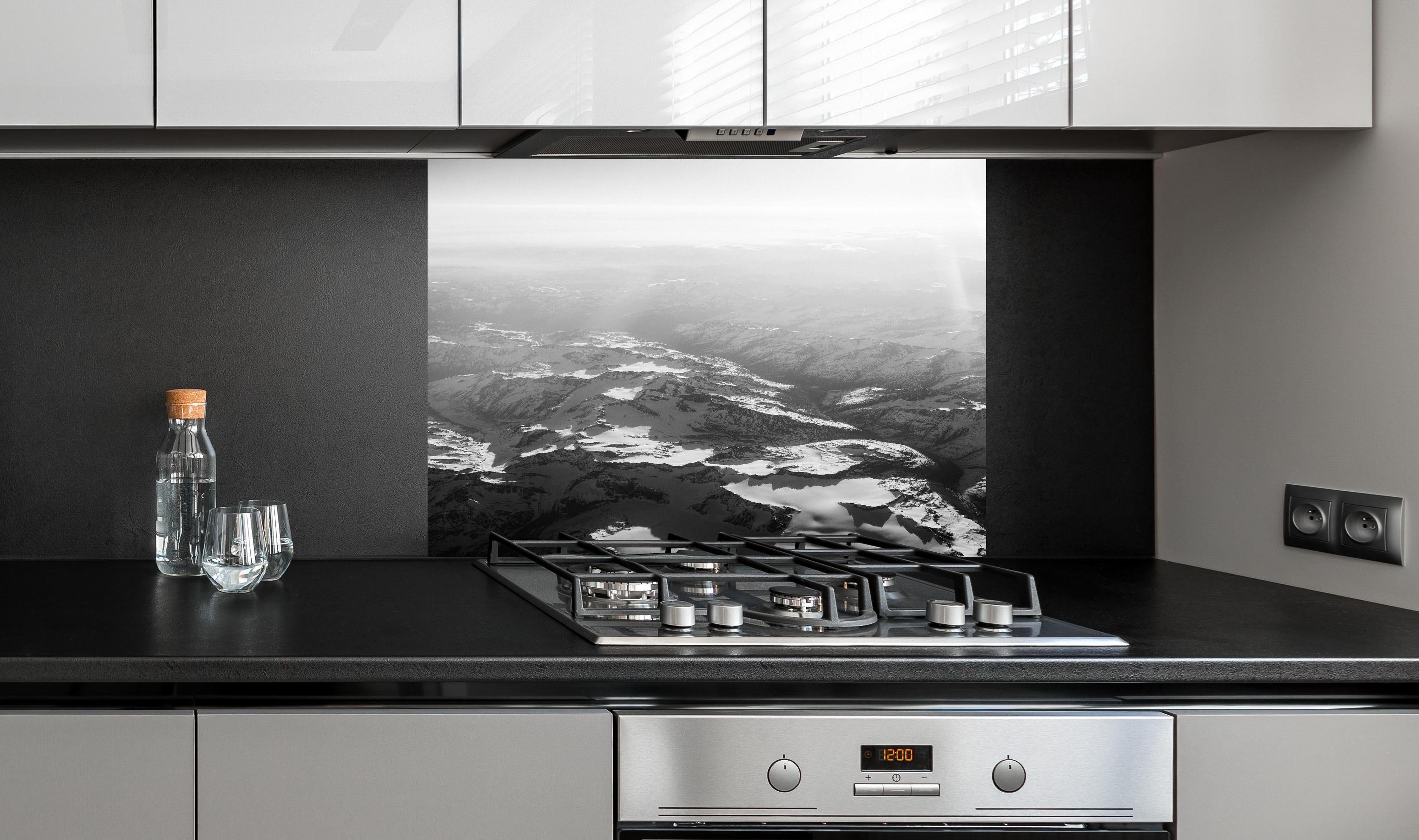 Spritzschutz - Schwarz-Weiß Fotografie Schneebedeckte Alpen hinter einem Cerankochfeld zwischen Holz-Kochutensilien
