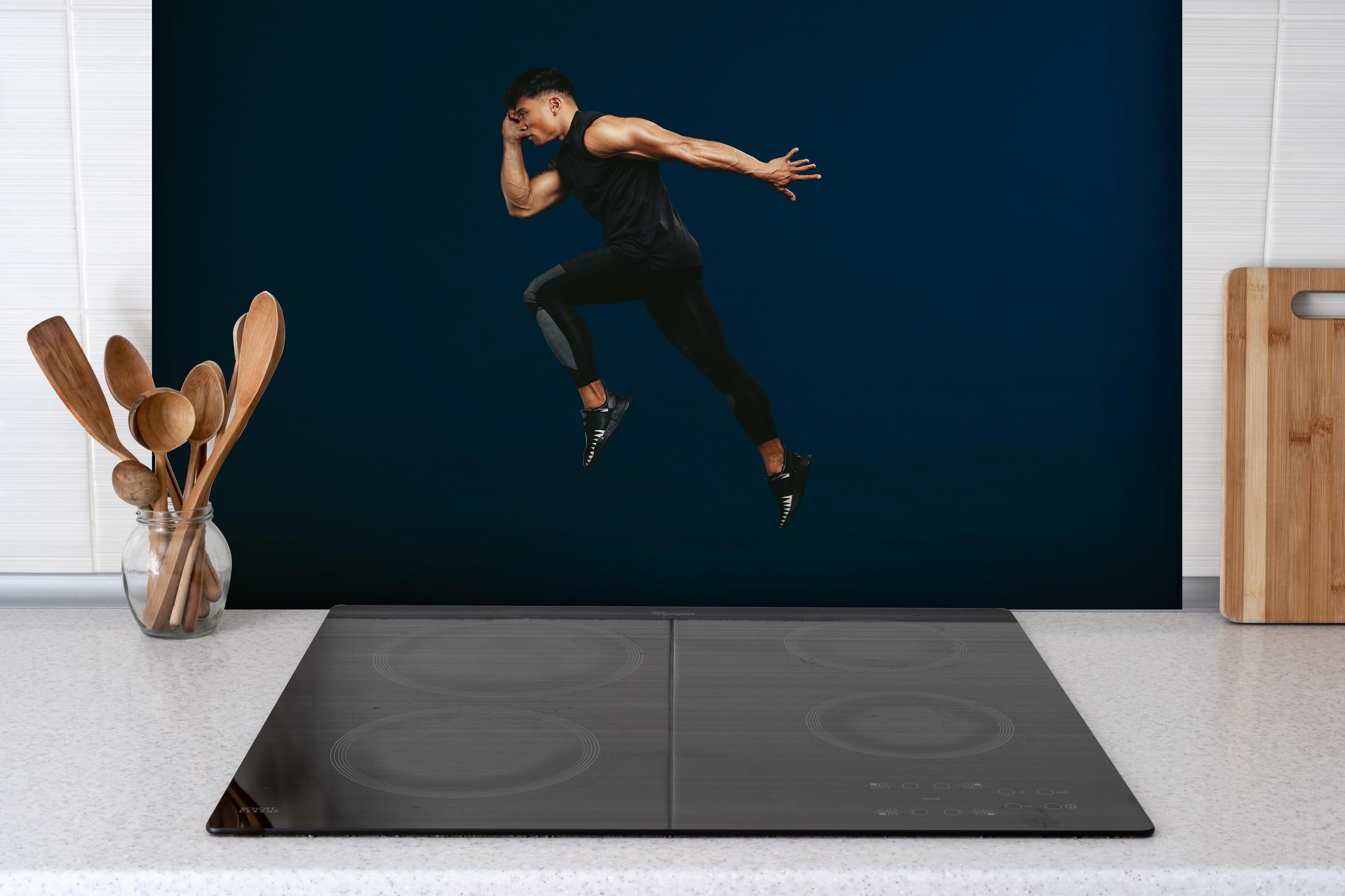 Spritzschutz - Sportler bei einem Sprung hinter einem Cerankochfeld zwischen Holz-Kochutensilien
