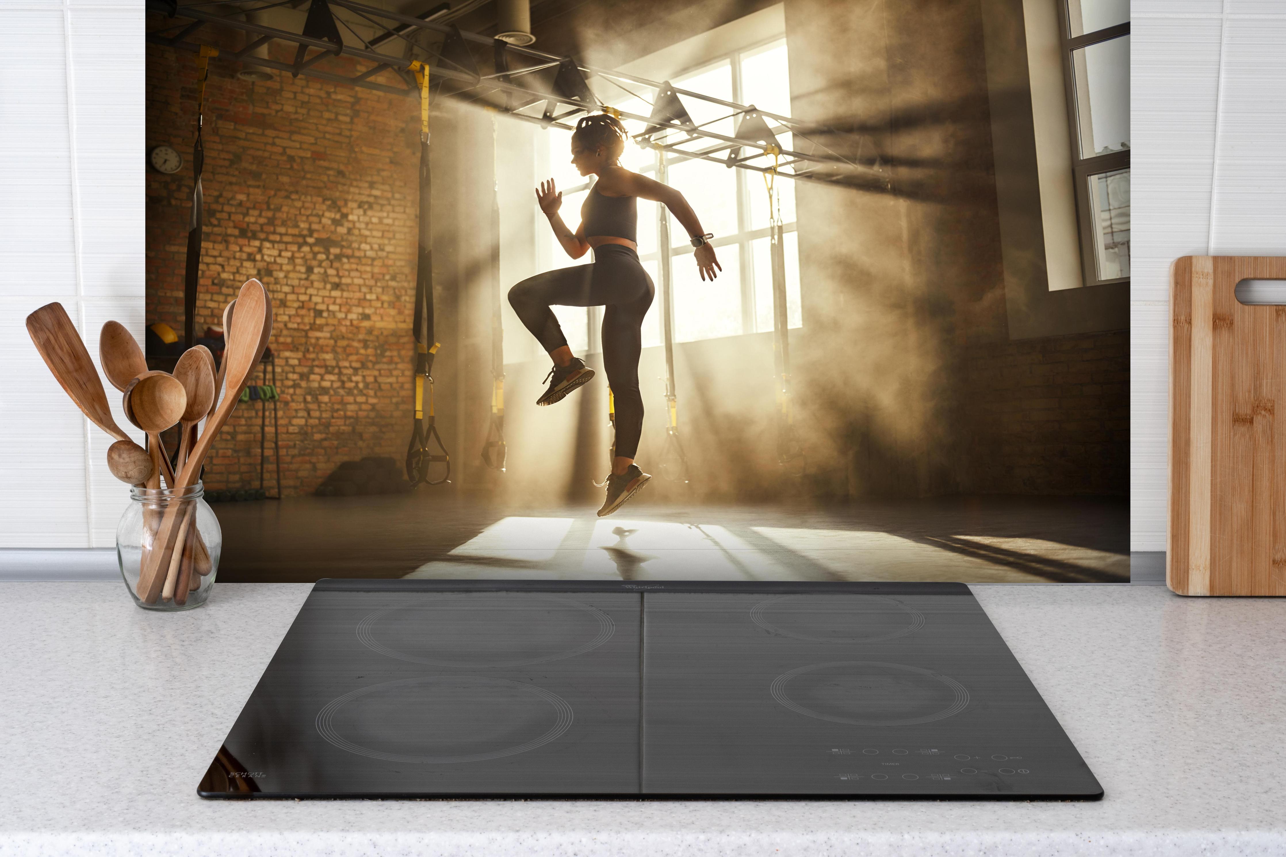 Spritzschutz - Sportlerin im Fitnesstudio hinter einem Cerankochfeld zwischen Holz-Kochutensilien
