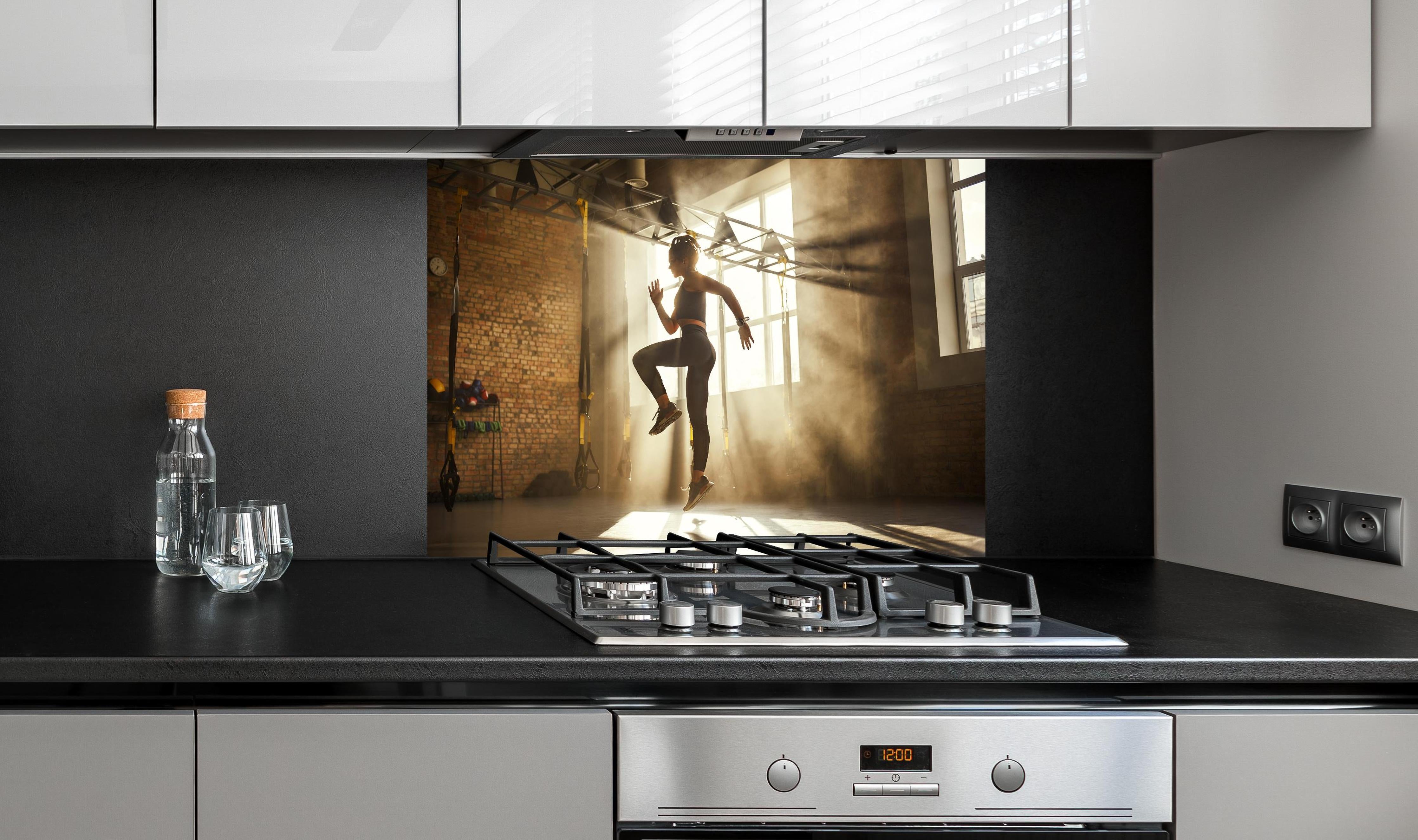 Spritzschutz - Sportlerin im Fitnesstudio hinter einem Cerankochfeld zwischen Holz-Kochutensilien
