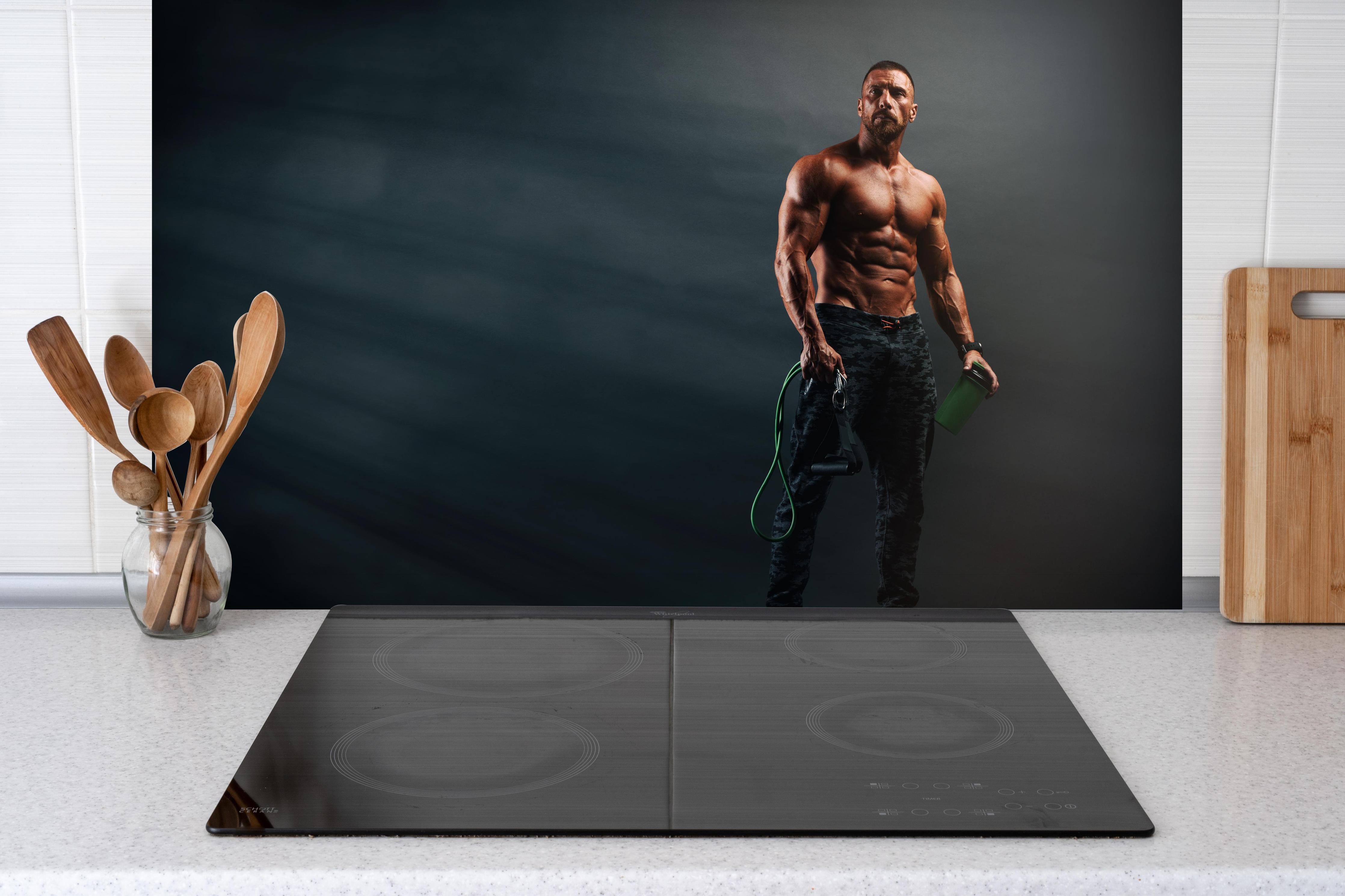 Spritzschutz - Bodybuilder - Training hinter einem Cerankochfeld zwischen Holz-Kochutensilien
