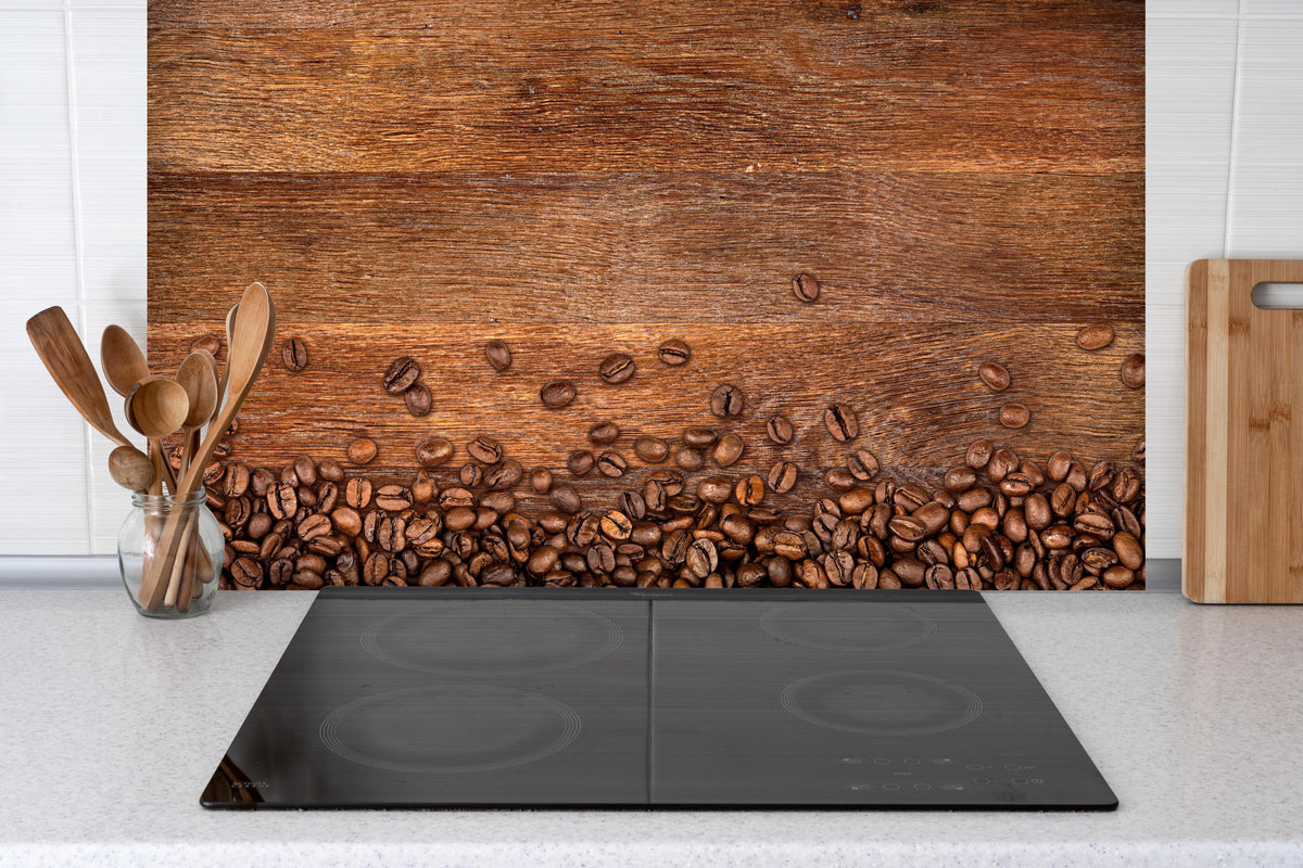 Spritzschutz - Kaffeebohnen auf Eichenholz hinter einem Cerankochfeld zwischen Holz-Kochutensilien
